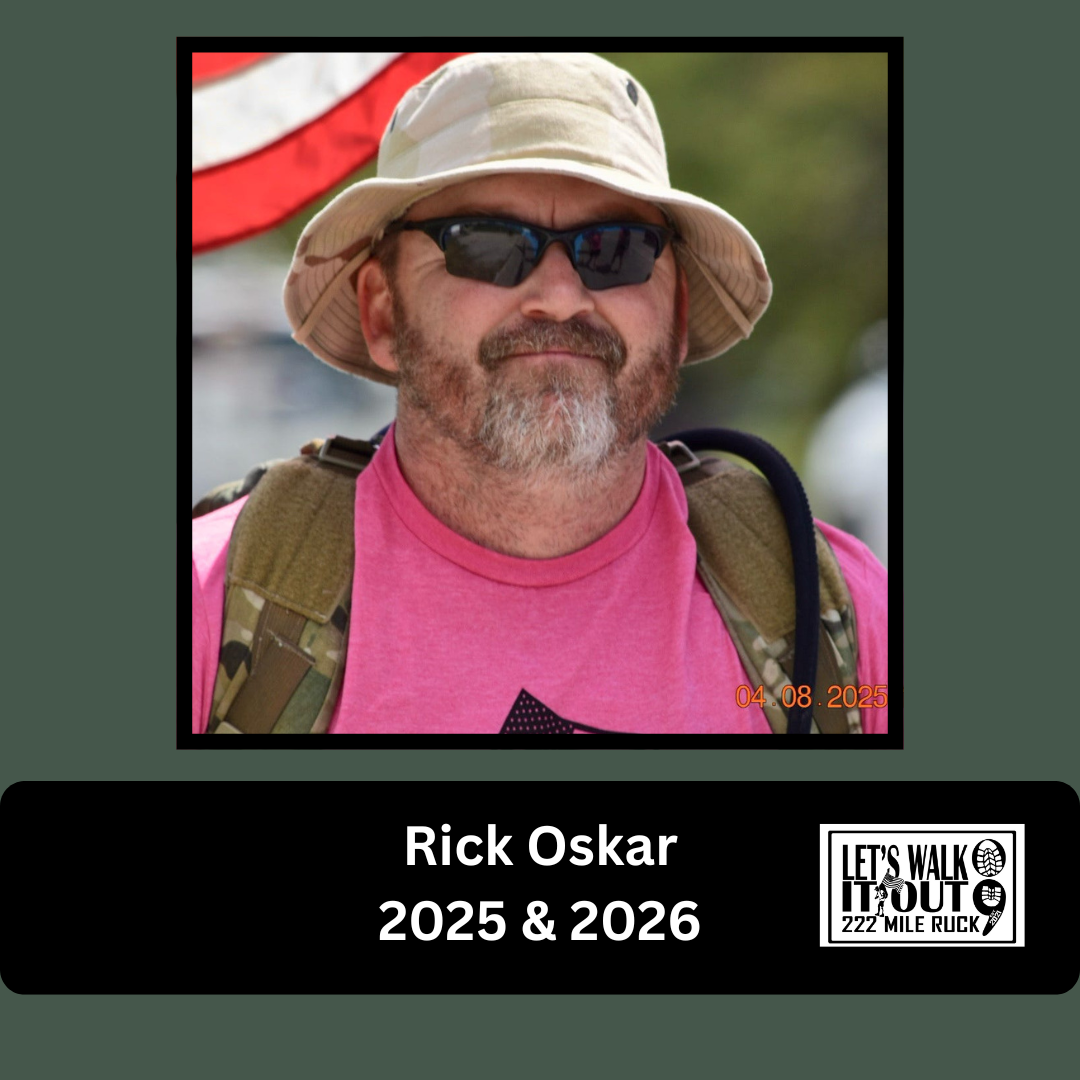 A man with a beard wearing a beige bucket hat, sunglasses, a pink shirt, and a backpack, standing outdoors with blurred background and an American flag in the background.