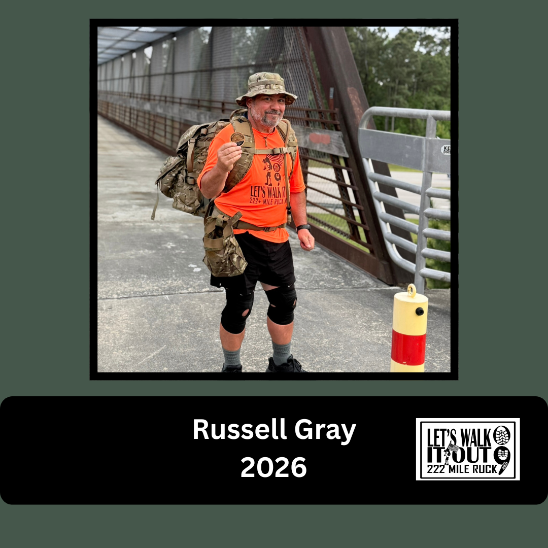 A man holding a medal, wearing a camouflage hat, orange T-shirt, black shorts with knee support, and carrying a large backpack, standing on a bridge with greenery in the background. He is smiling and posing for the camera.