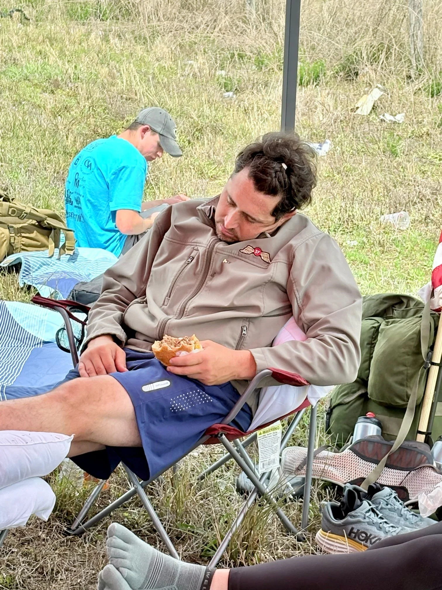 A man resting in a camping chair outdoors, holding a sandwich with his eyes closed. He is in a field with grass, surrounded by camping gear and shoes, with another person sitting nearby in the background.