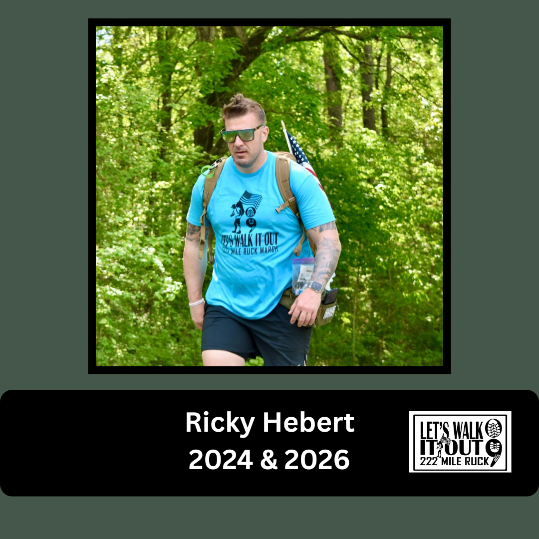 A man with sunglasses and tattoos wearing a blue t-shirt and black shorts walking in a green forested area, carrying a backpack with an American flag patch and a small pouch attached to his waist.