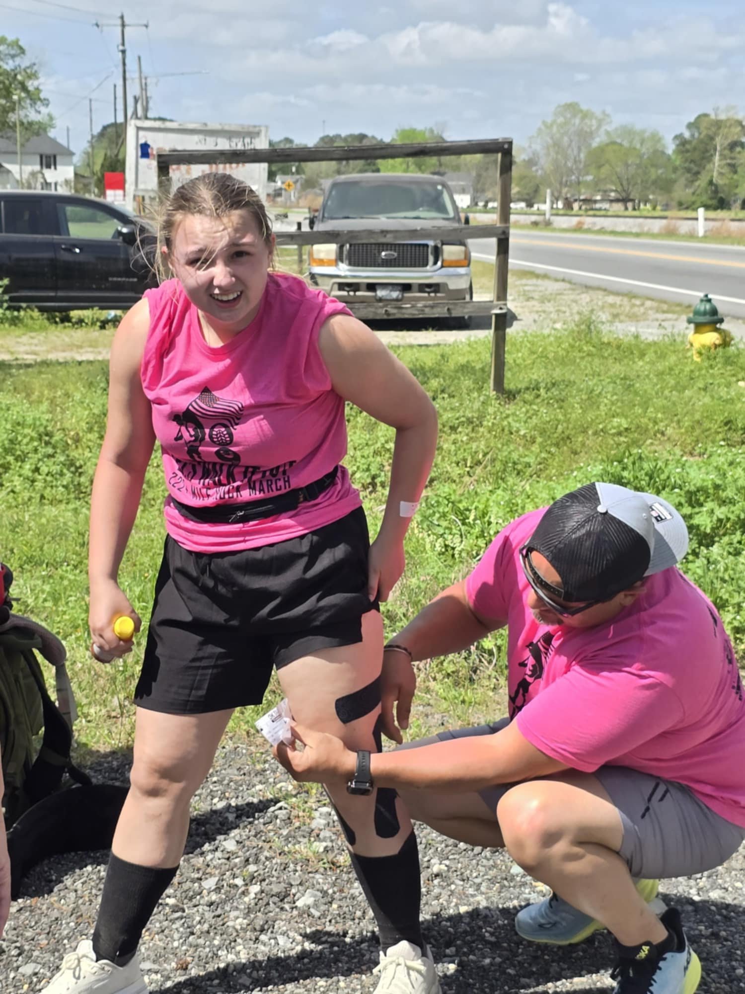 A young girl with a bandage on her arm is wincing as a man applies a band-aid to her knee, both wearing pink shirts, in an outdoor grassy area near a road with vehicles in the background.