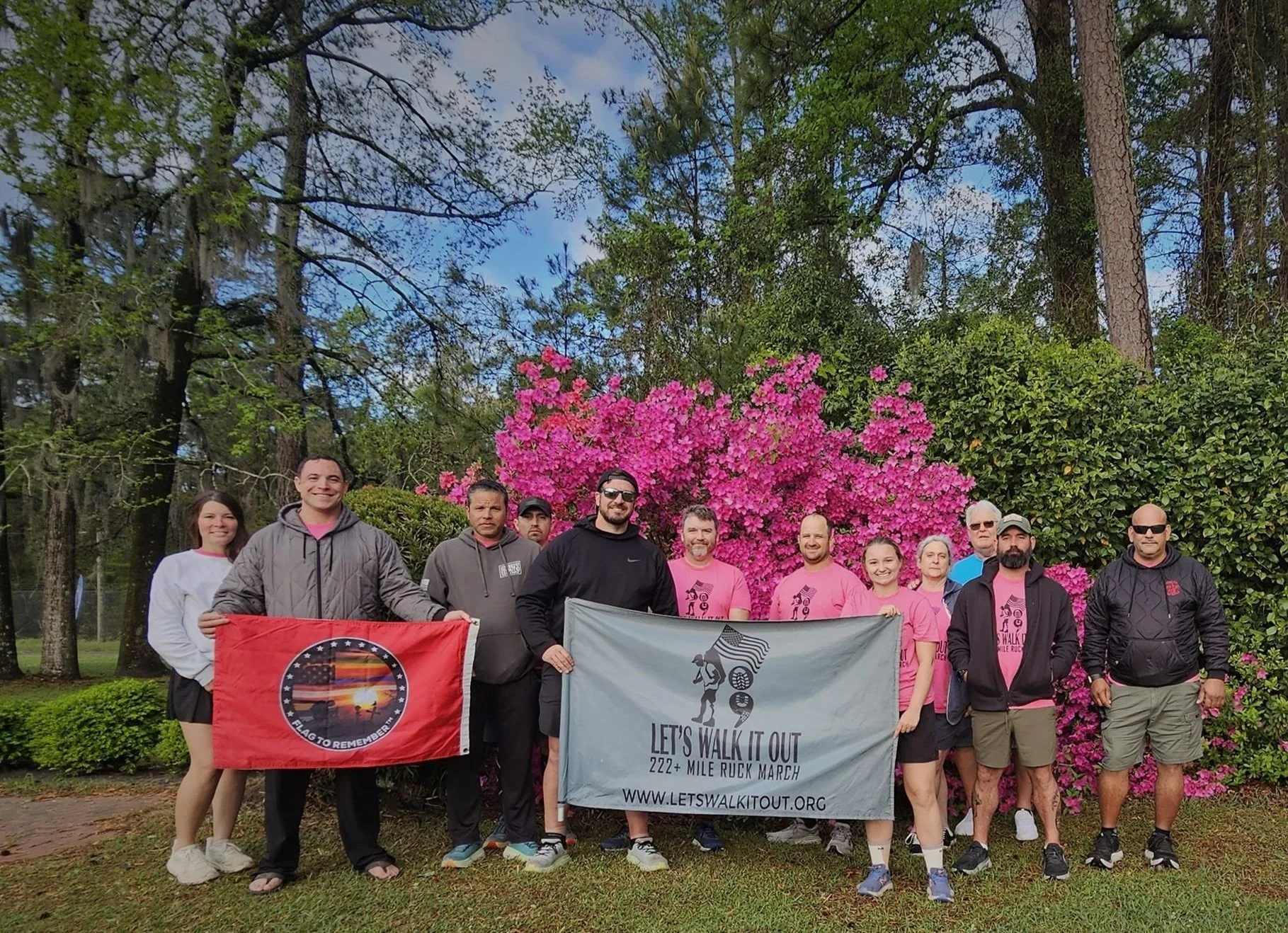 Group of people holding flags in front of pink blooming bush, with trees and greenery in the background.
