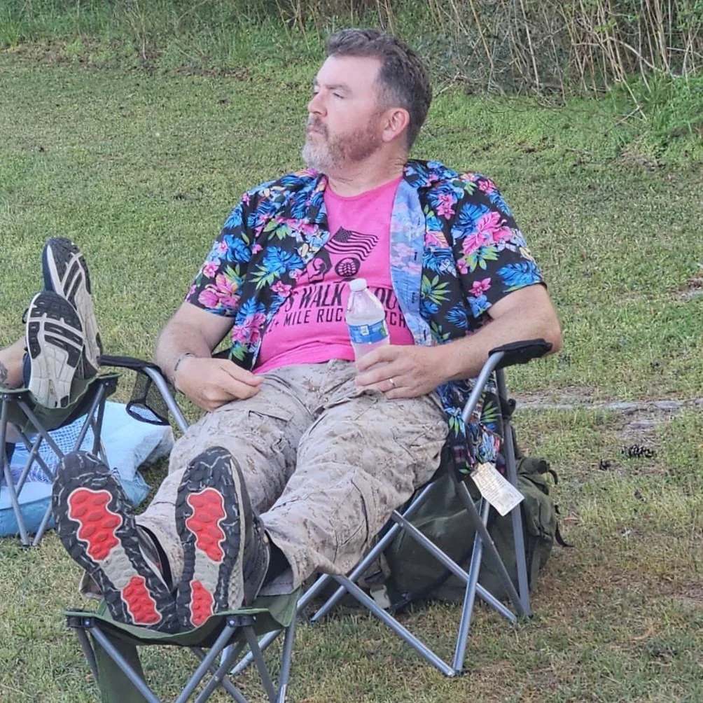A man with a beard sitting outdoors on a camping chair, wearing a colorful Hawaiian shirt, camouflage cargo shorts, and holding a water bottle, with his eyes closed and leaning back against a grassy area with bushes.