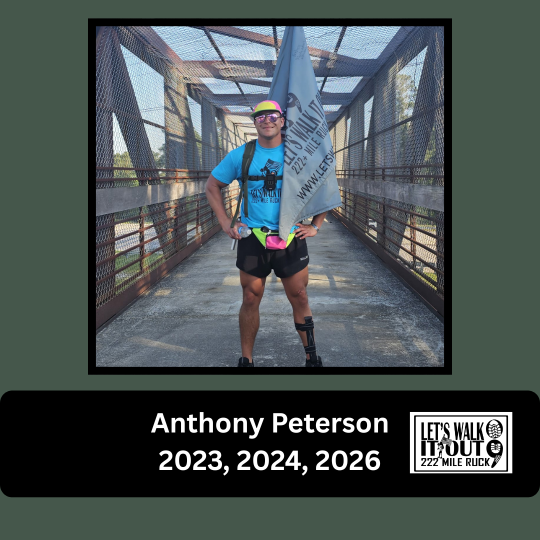 Man standing on a bridge holding a flag, wearing a blue shirt, black shorts, sunglasses, a visor, and a knee brace during a running event.