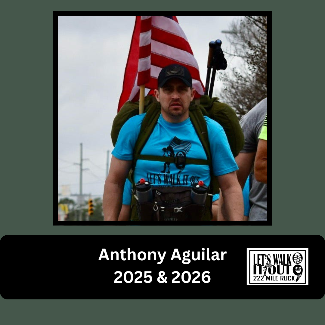 A man hiking outdoors carrying a backpack, American flag, and gear, wearing a blue t-shirt with a logo and text, with cloudy sky and power lines in the background.