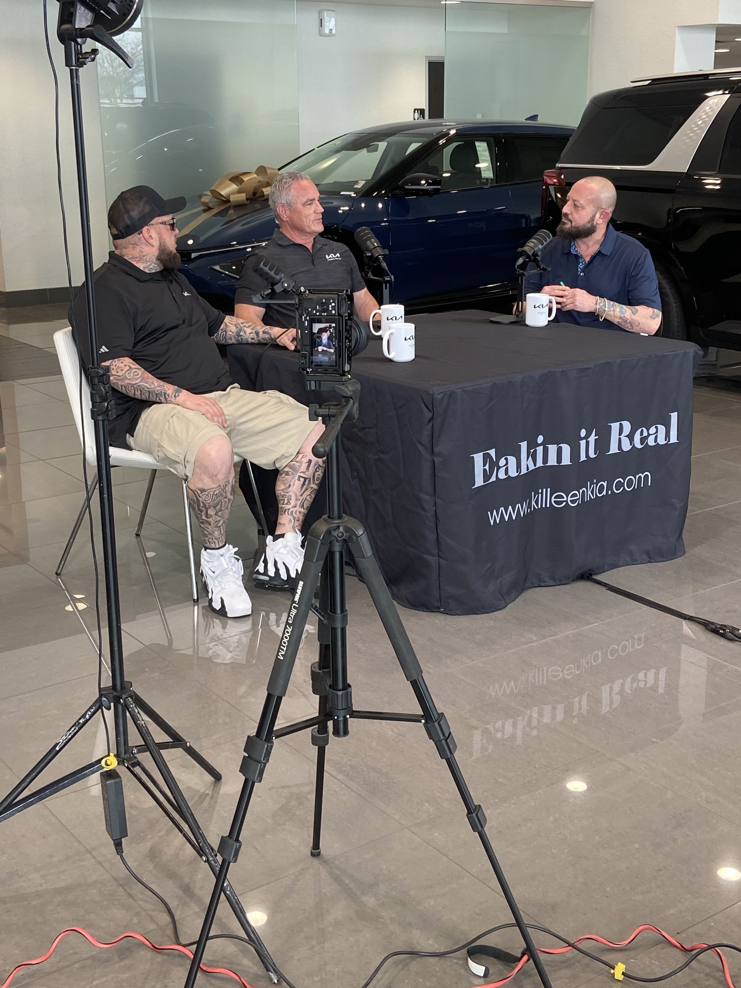 Three men sitting at a table with microphones, recording a podcast or interview, in a showroom with cars in the background.
