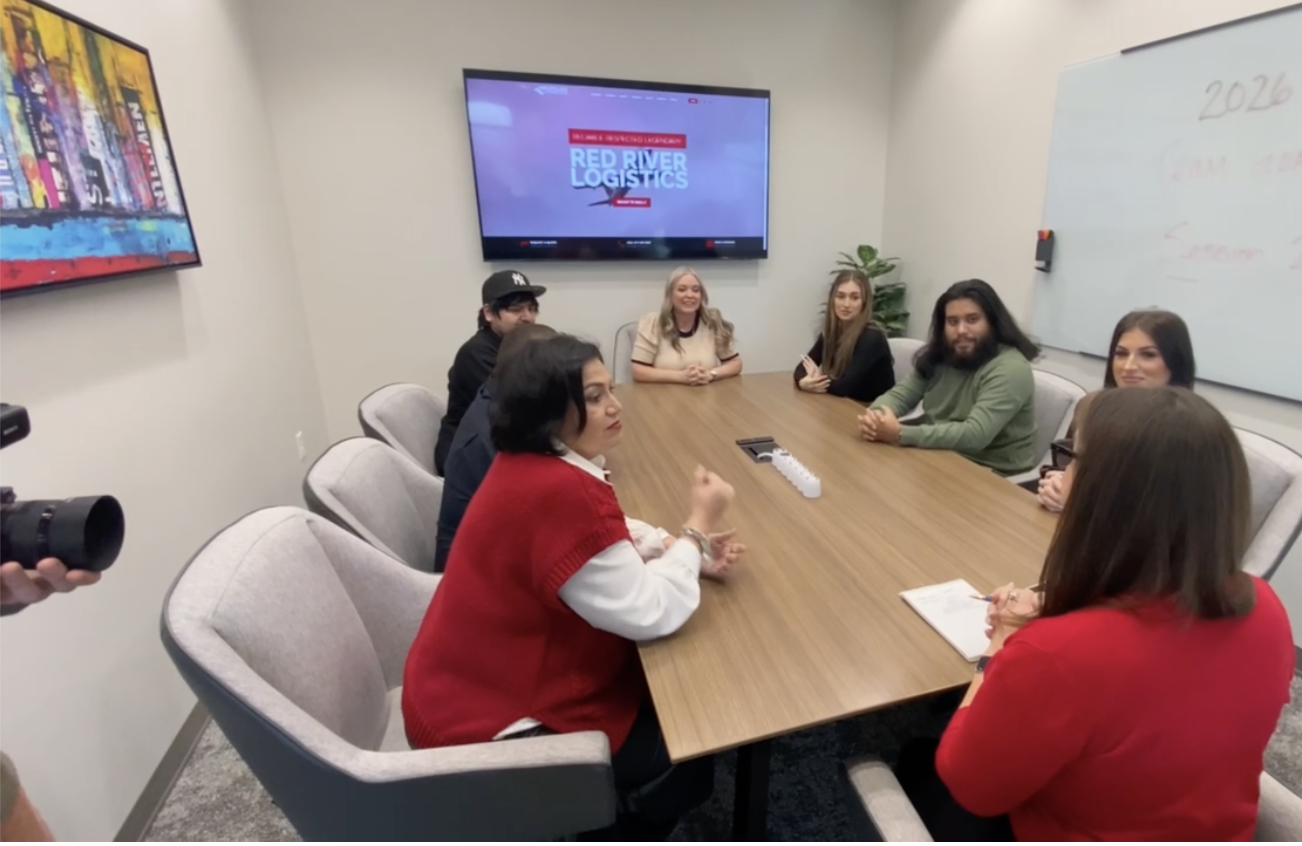 People attending a meeting around a conference table in a modern office, with a whiteboard and a television screen displaying 'Red River Logistics' in the background.