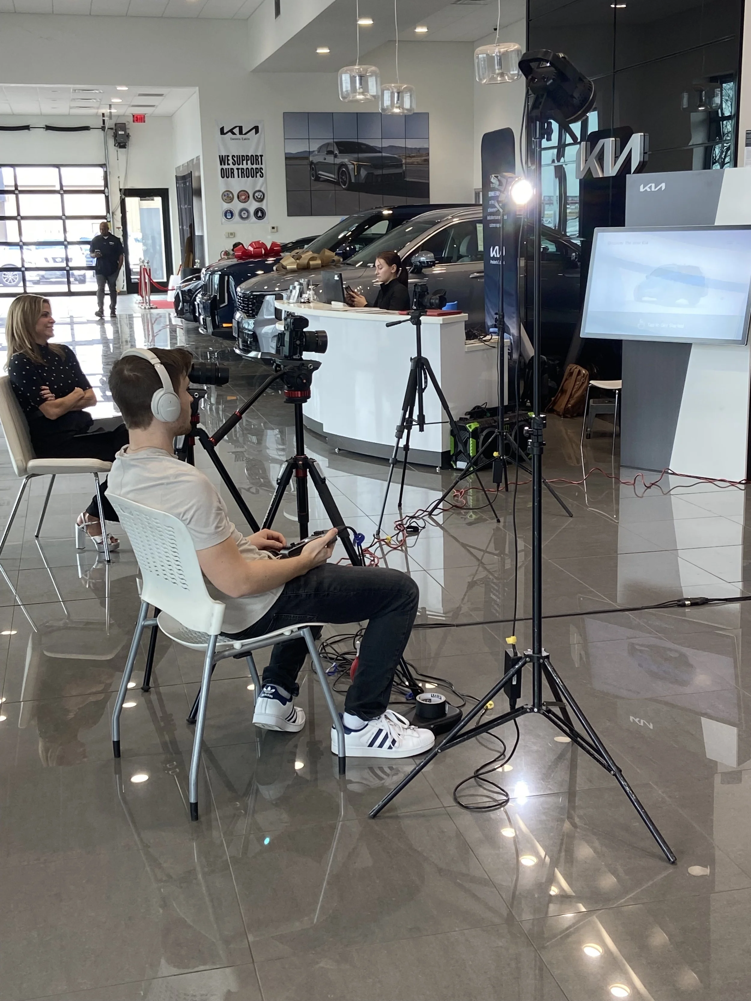 Recording crew setup in a car dealership showroom with a camera and lighting equipment. Two women are seated, one being interviewed or filmed, with cars and dealership signage in the background.