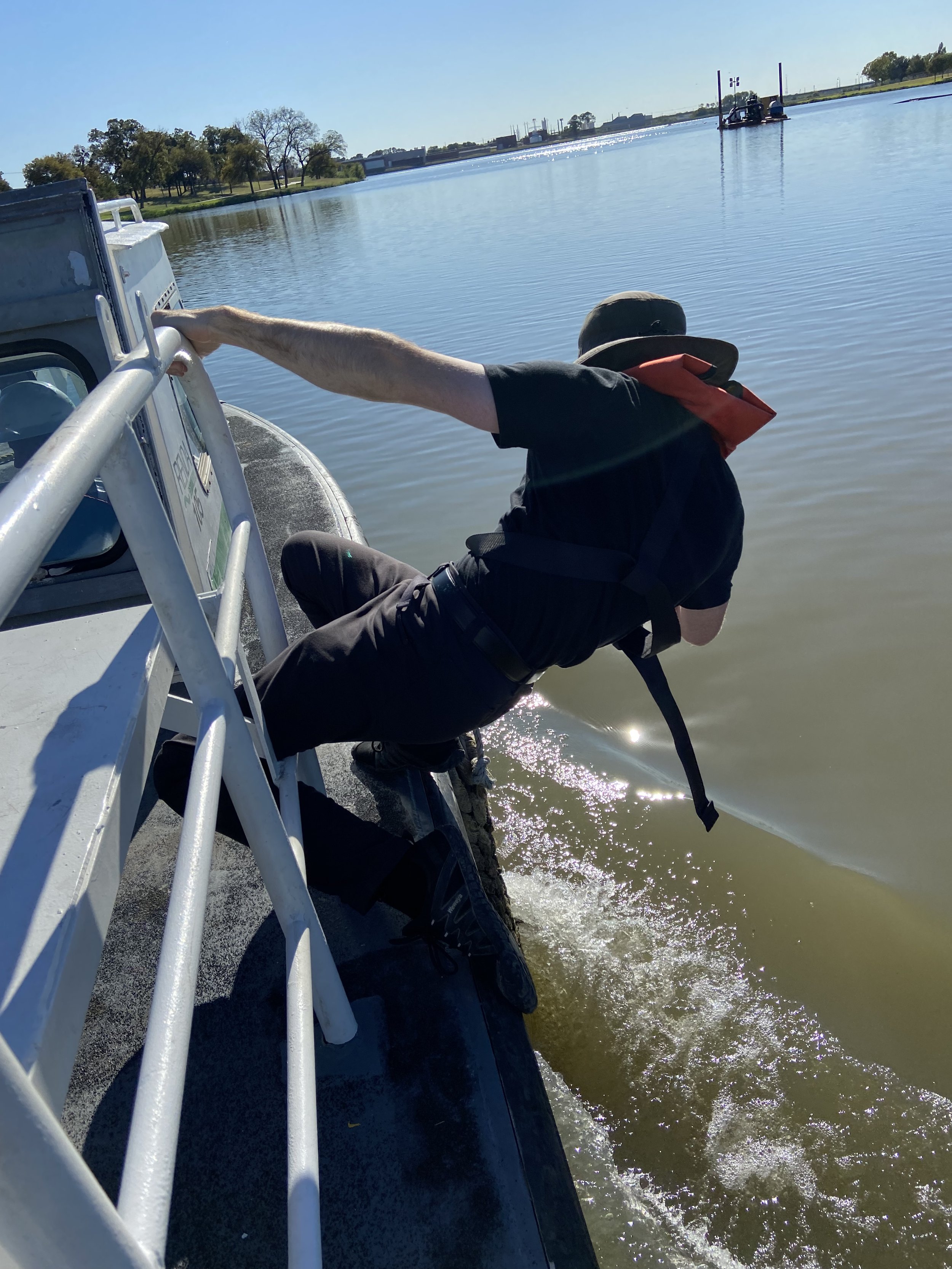 A person leaning over the side of a boat, holding onto a railing, with water and a shoreline with trees in the background.