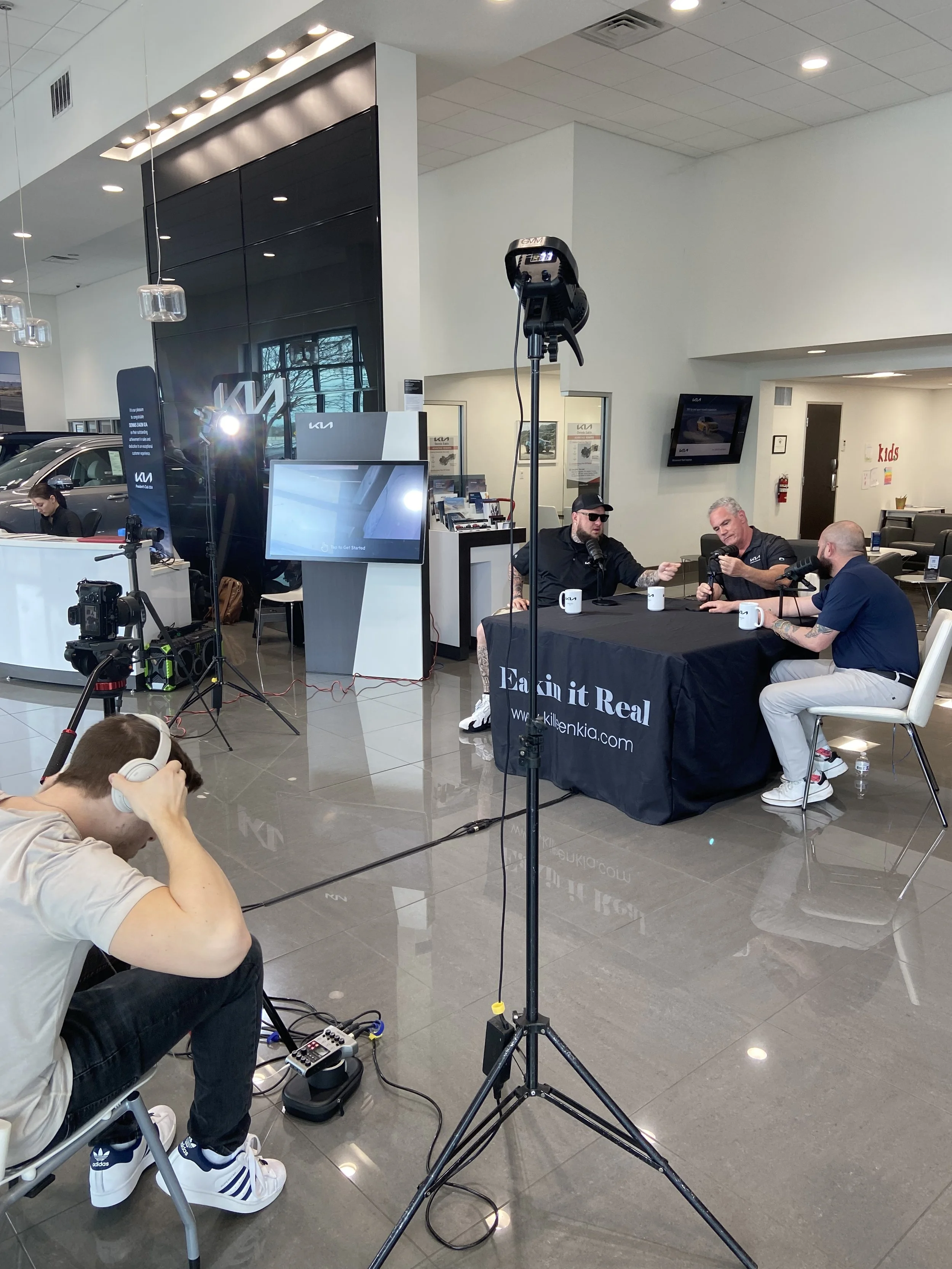 A podcast recording session with three men sitting at a table with microphones and mugs, inside a modern showroom with cars and a large wall display in the background.