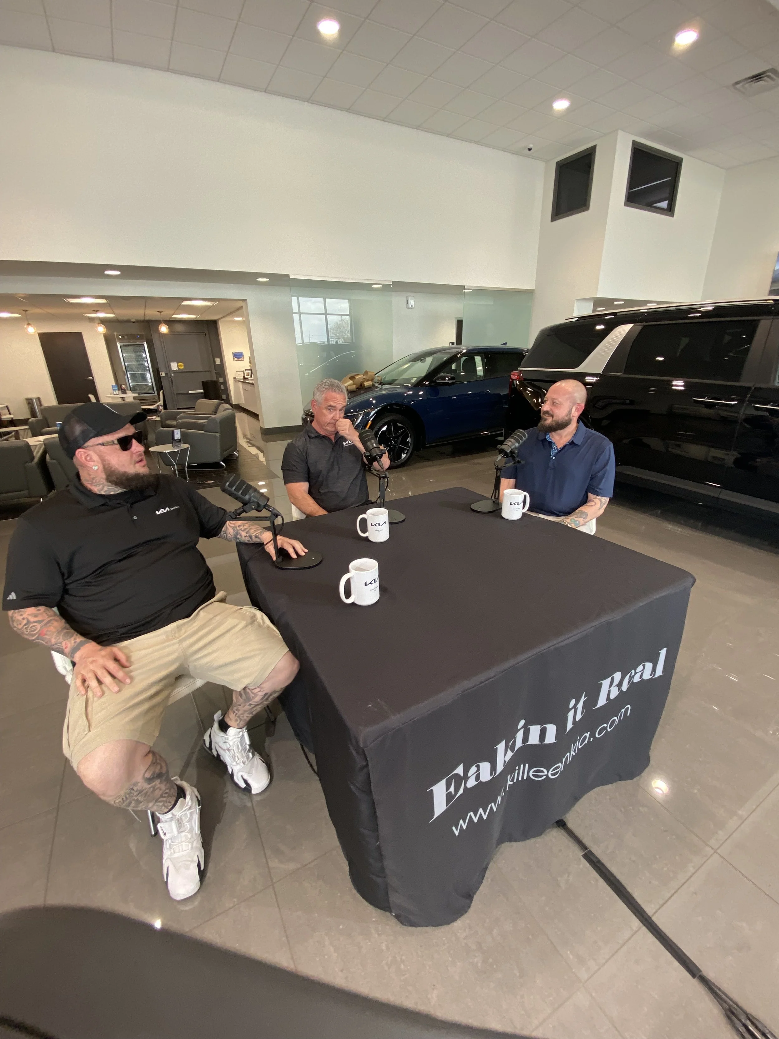 Three men participating in a podcast recording at a dealership. They are seated at a black covered table with microphones and coffee mugs. Cars are visible in the background inside the showroom.