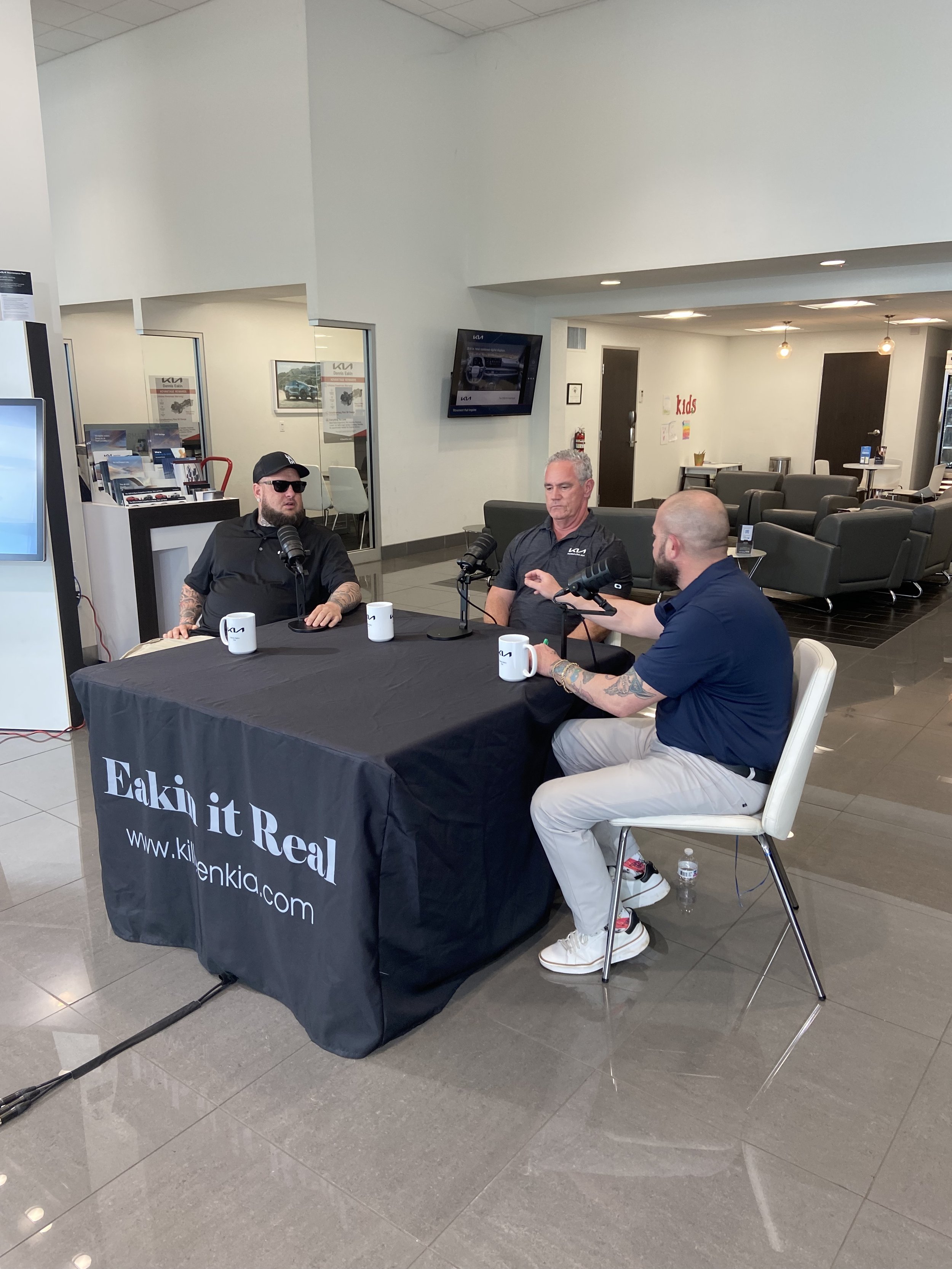 Three men sitting at a table with microphones, recording a podcast or interview inside a car dealership, with a sign that reads 'Eakim it Real' on the tablecloth.