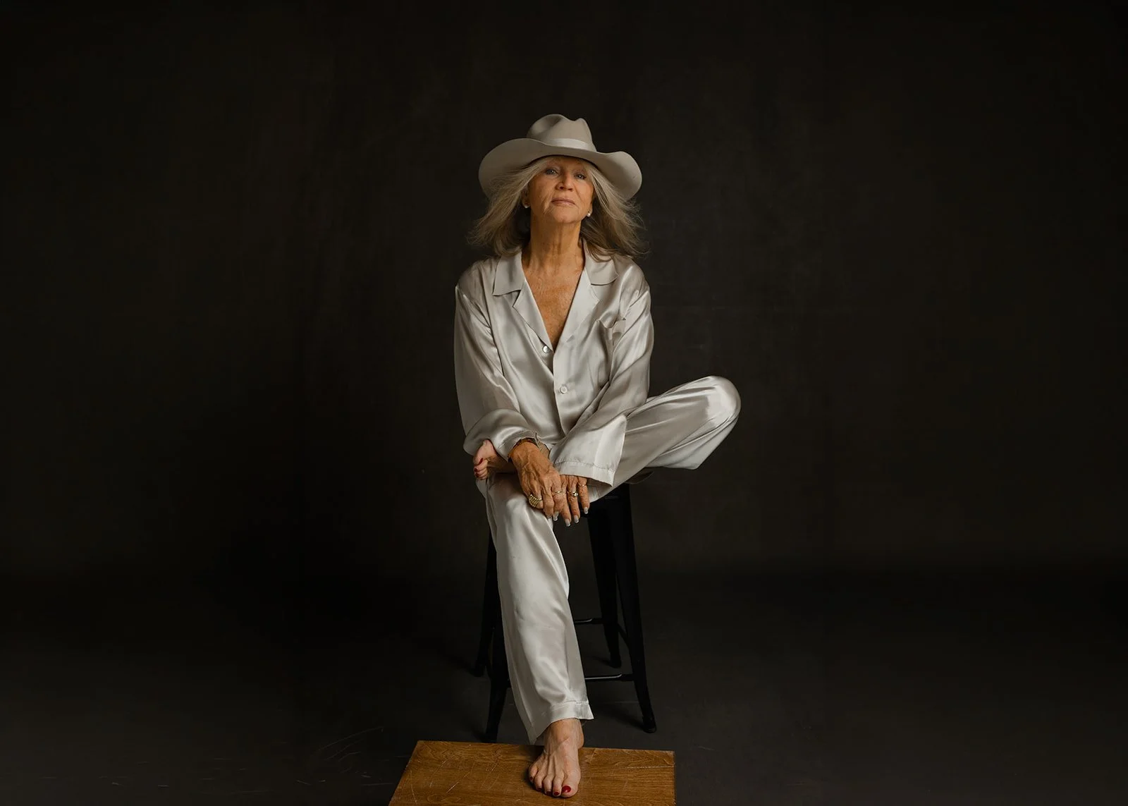 Leigh Kilton-Smith sits barefoot on a stool wearing a cream satin suit and wide-brimmed hat in a thoughtful portrait created for the 50 Women Project by Christine Harris.