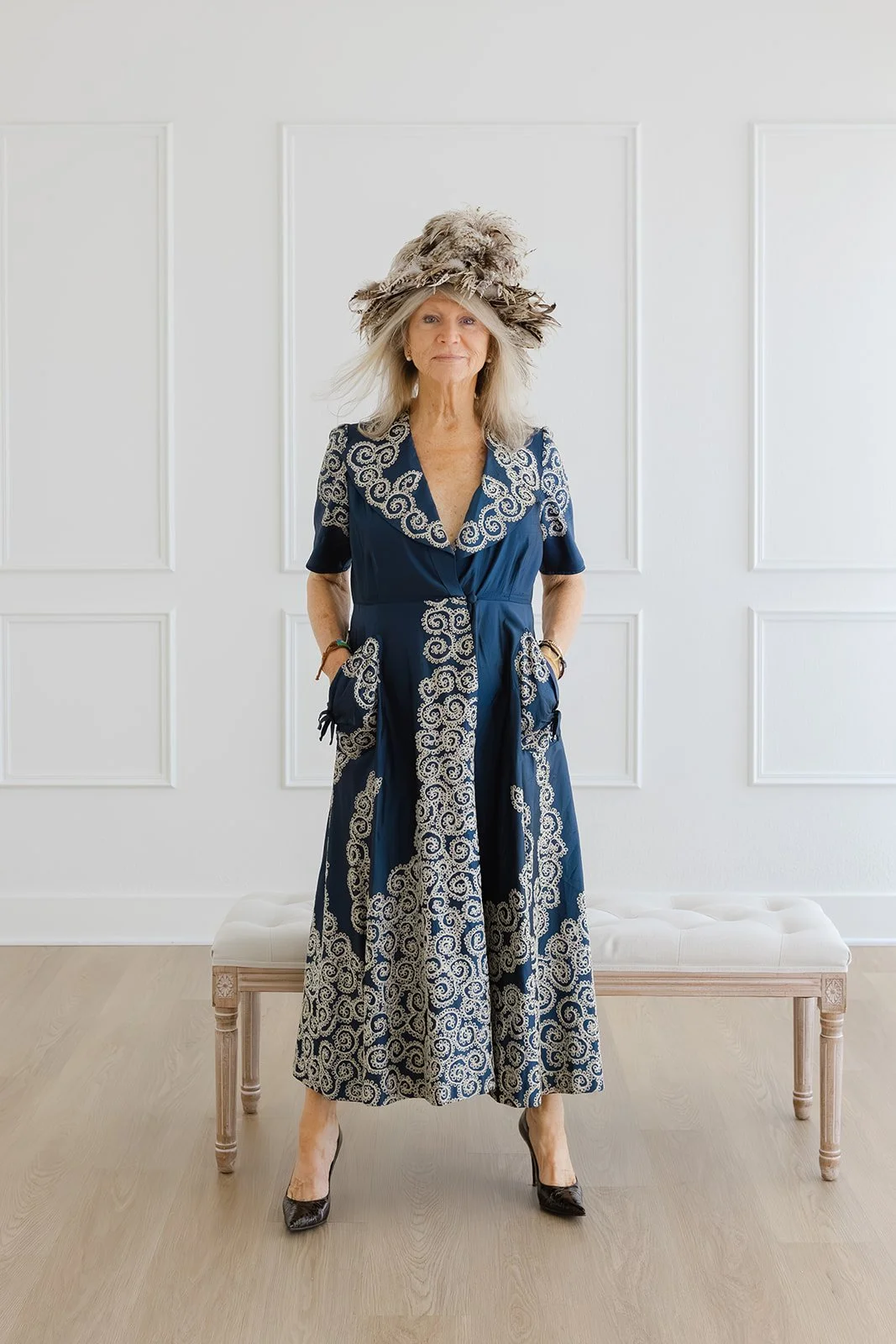 Leigh Kilton-Smith stands confidently in a blue patterned dress and feathered hat beside a vintage bench during her 50 Women Project portrait session by Christine Harris.