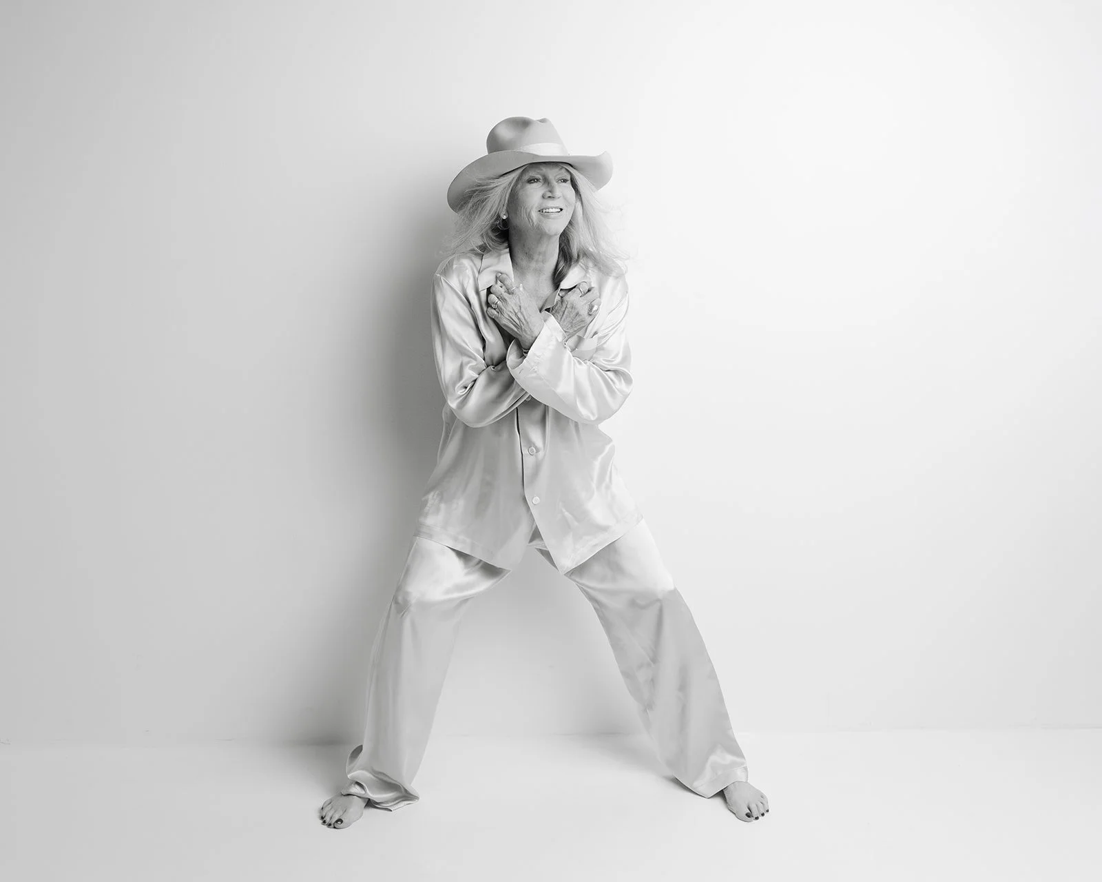 Black-and-white portrait of Leigh Kilton-Smith standing barefoot in a cream satin suit and wide-brimmed hat, smiling with arms crossed over her chest during her 50 Women Project session by Christine Harris.