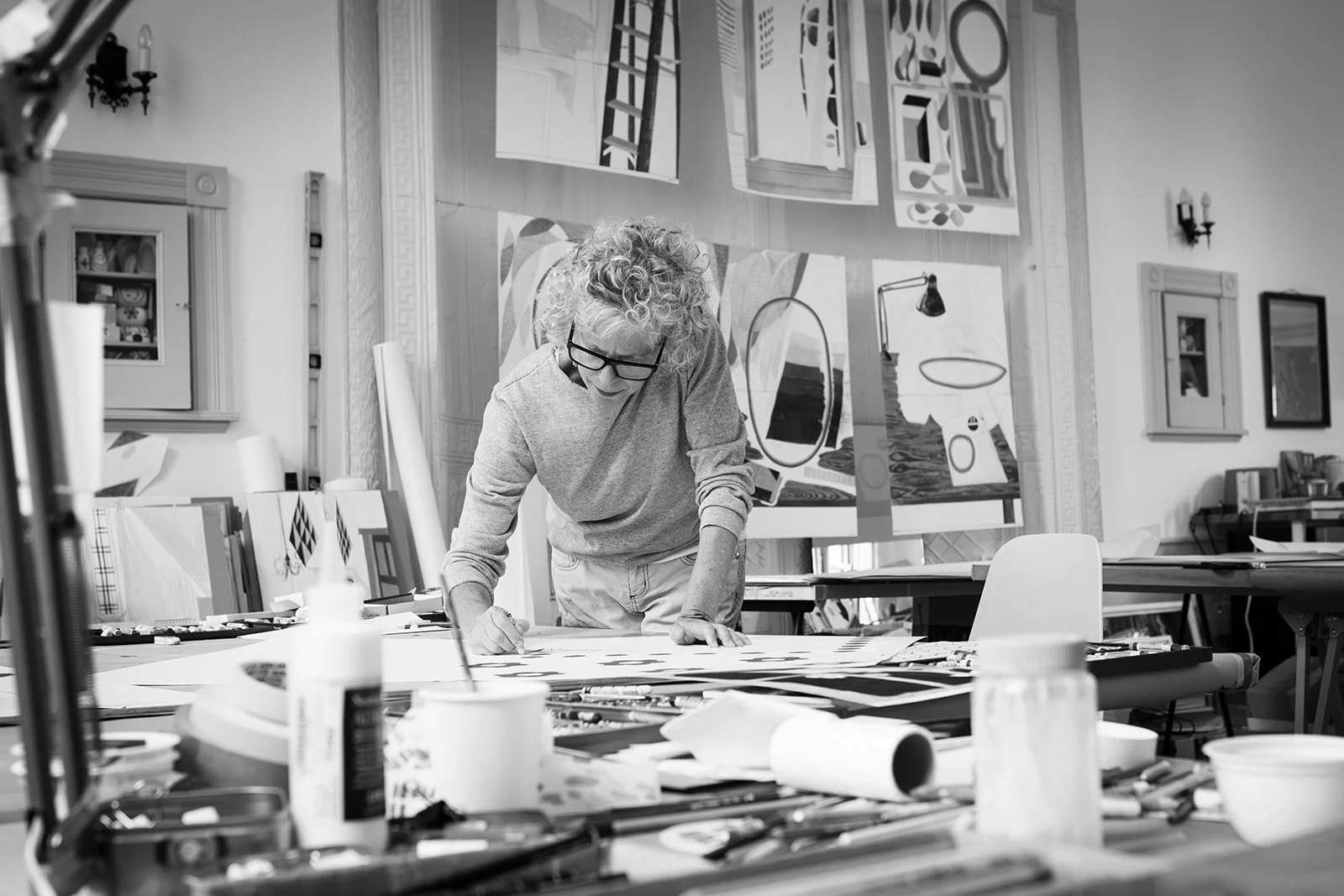 A black and white portrait of artist Michelle Weinberg leaning over her worktable in her Sharon Springs studio, surrounded by her paintings and tools, photographed by Christine Harris.