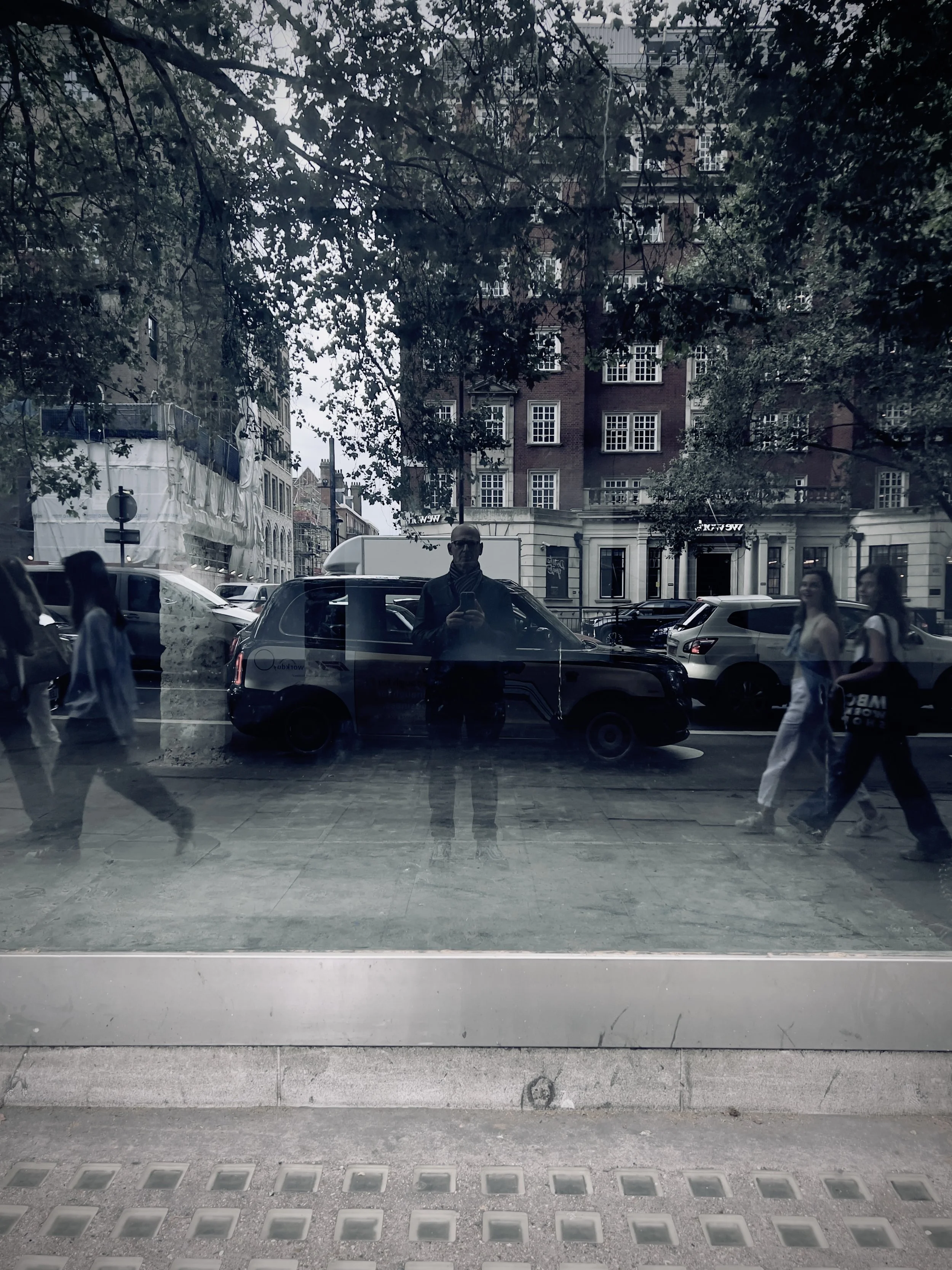 City street scene reflected in a glass window, showing pedestrians walking, cars parked along the street, and a man taking a photo with his phone.