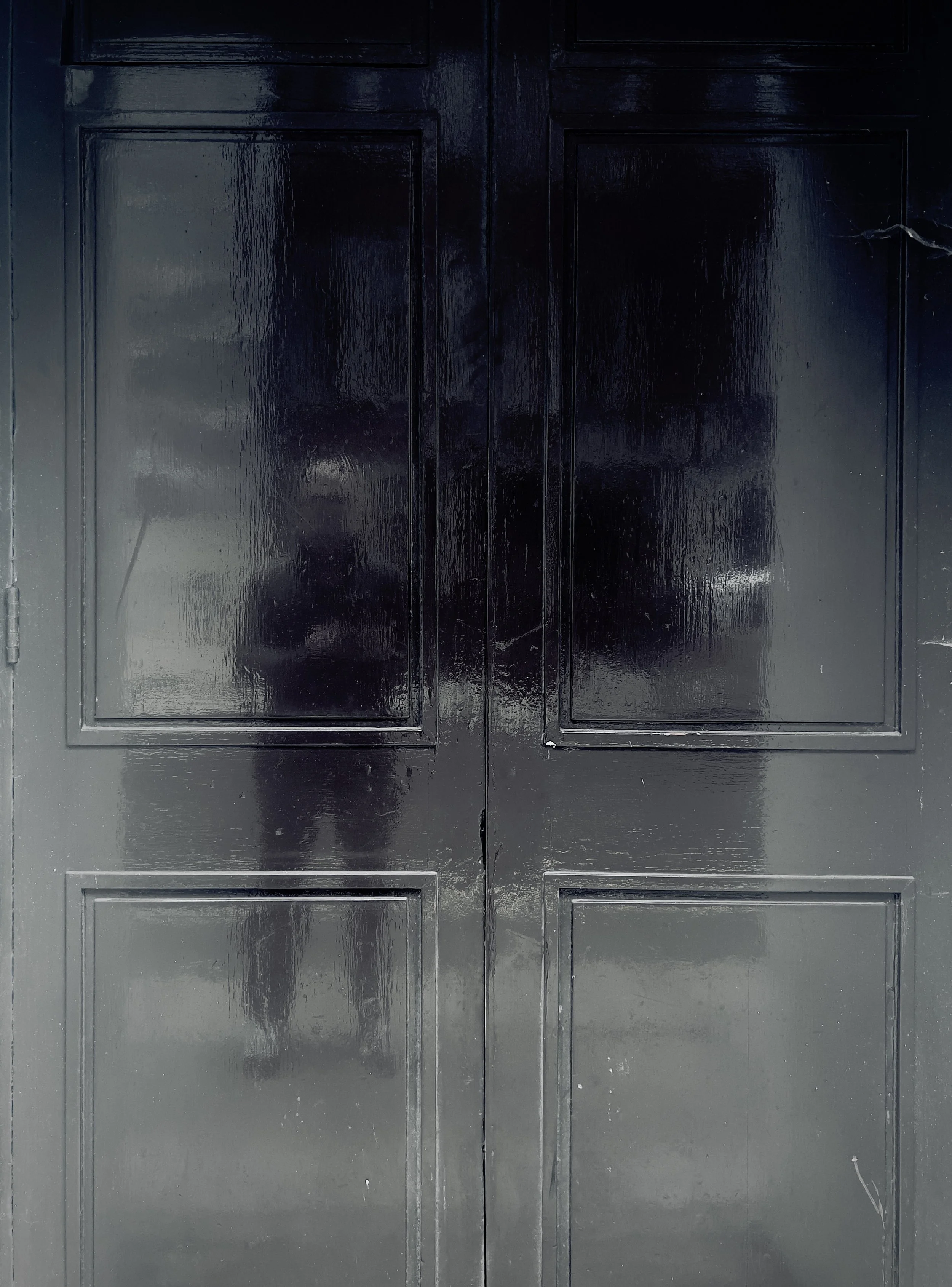 Black double doors with a glossy finish, featuring four recessed panels each, and a small area where the paint appears chipped or scratched.