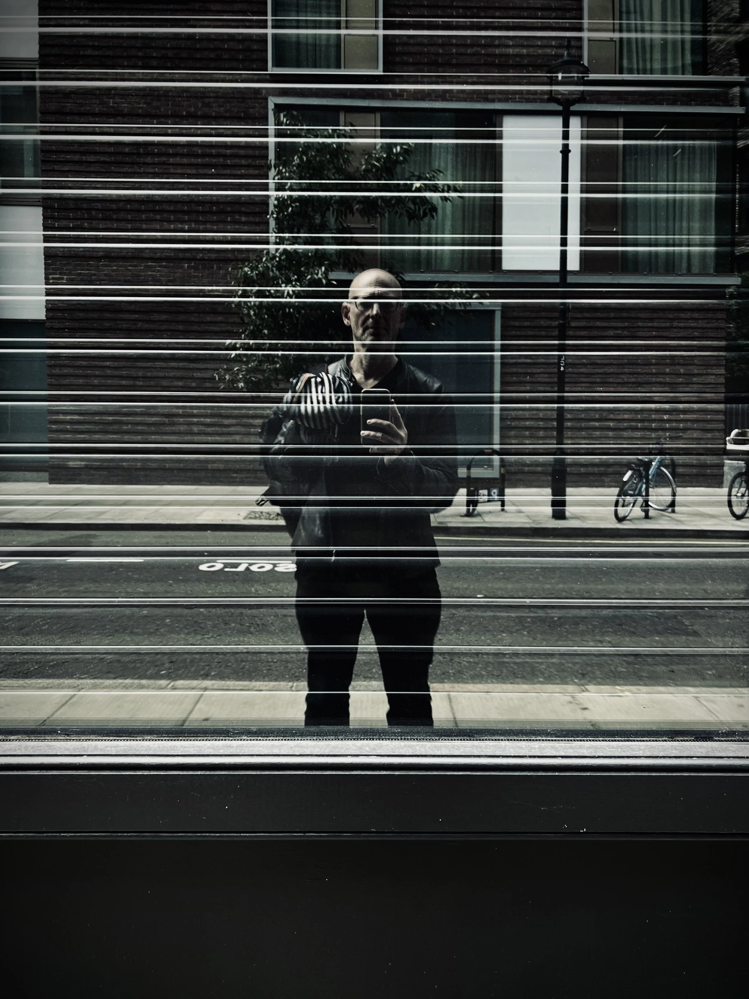 Self-portrait of a man taking a photo of his reflection in a glass window with horizontal blinds, with a city street and bicycles in the background.