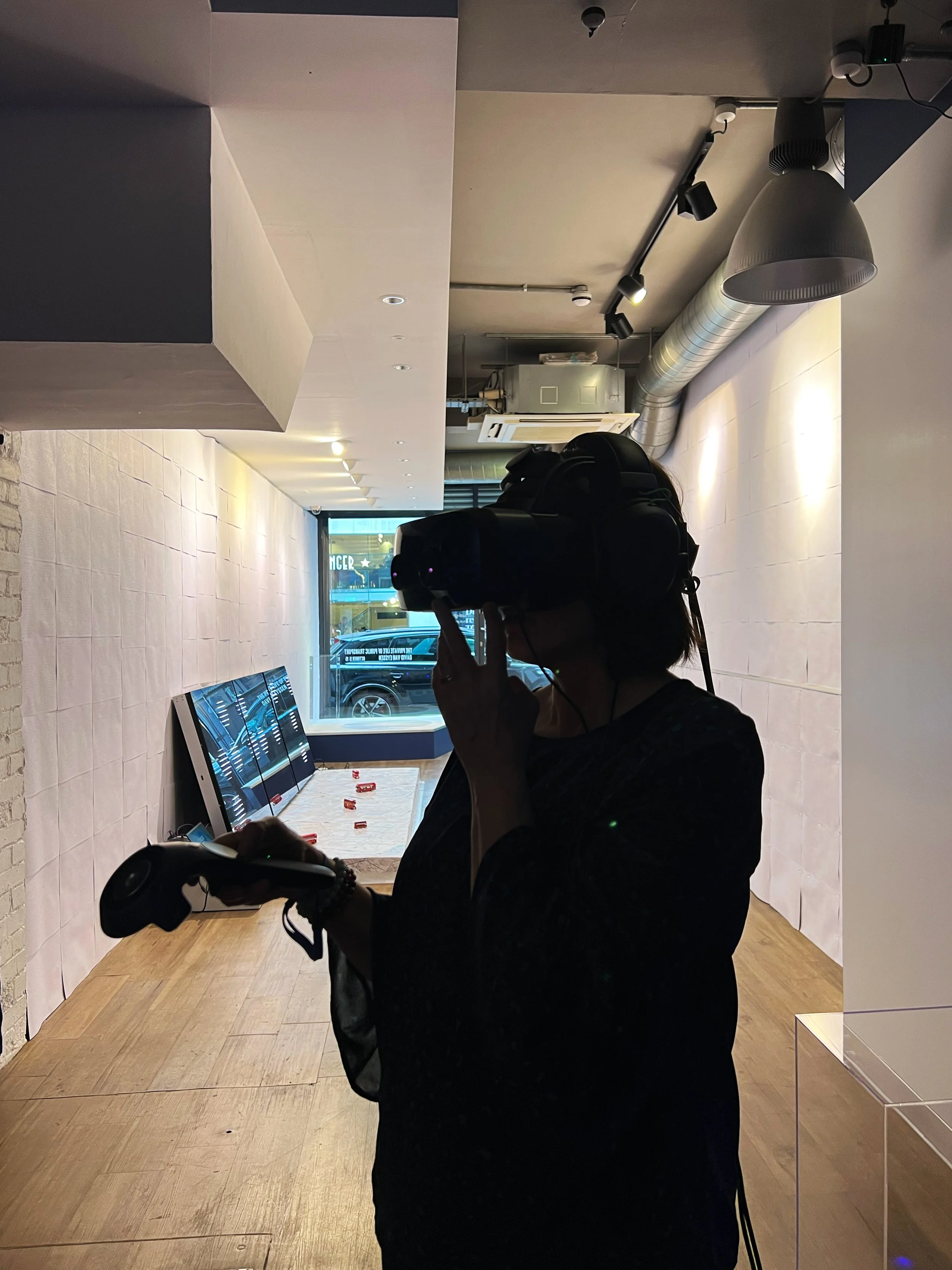Person using a virtual reality headset and controller in a modern room with large windows and wall displays.