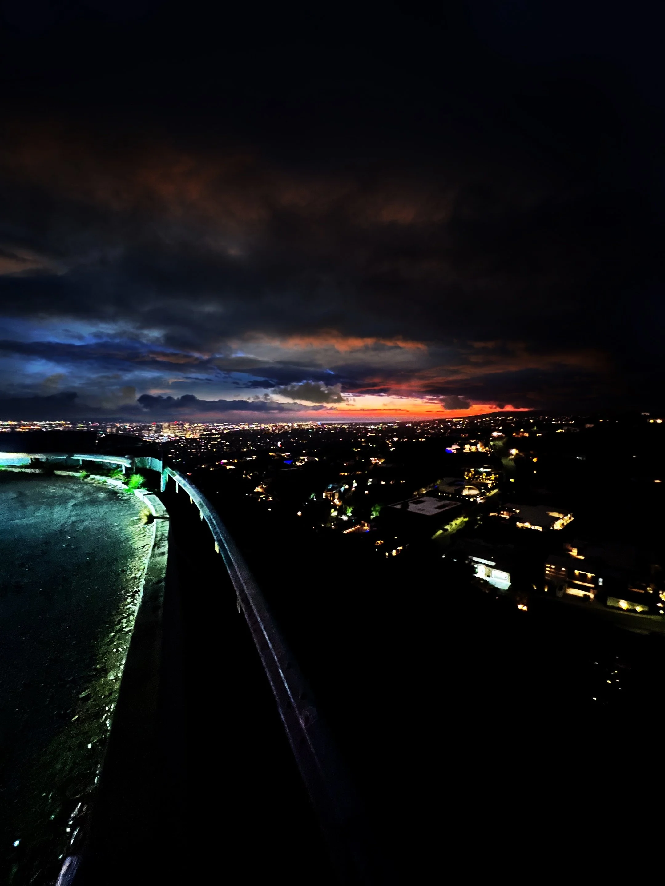 Scenic view of a city at night with illuminated buildings and dramatic clouds at sunset, visible from a roadside viewpoint.