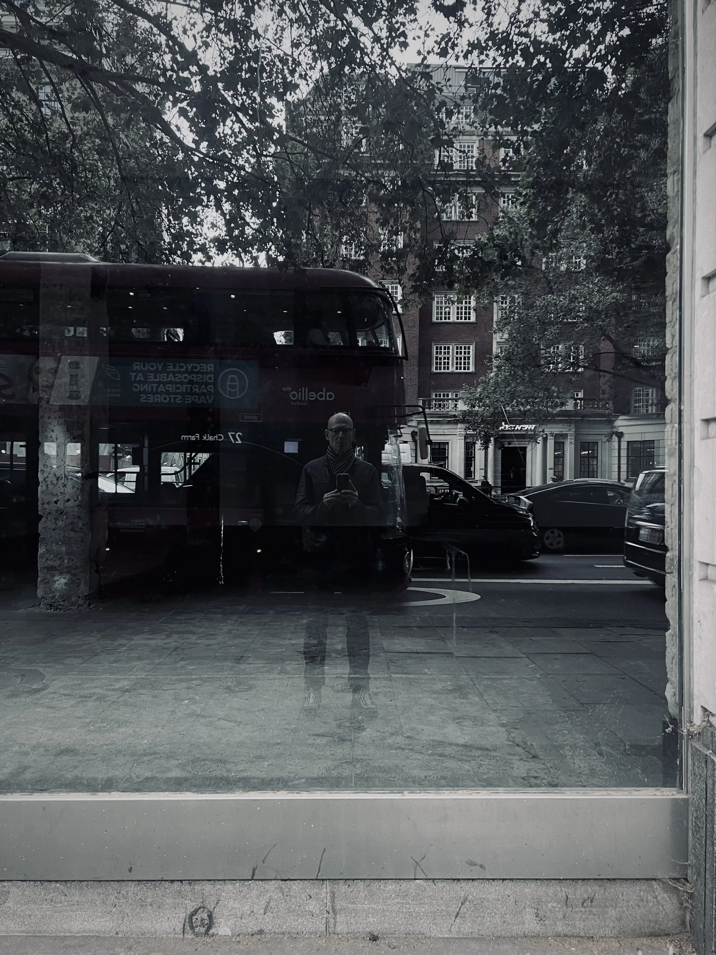 Reflection of a person taking a photo in a window with a red double-decker bus, trees, and buildings visible outside.