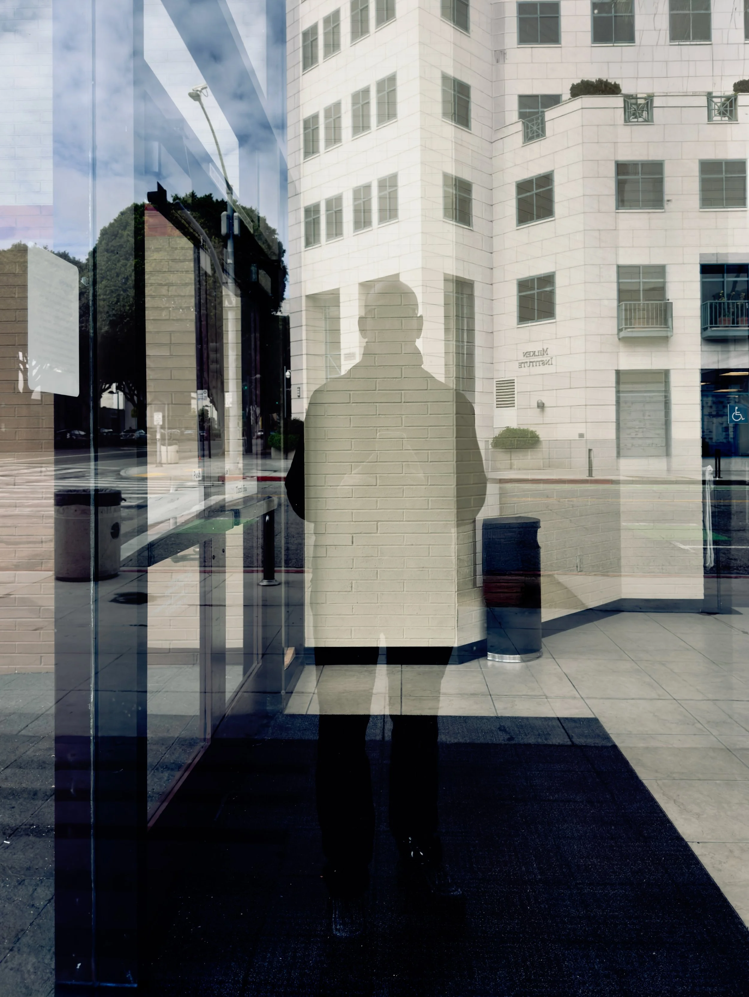 A reflection of a person standing outside a building, visible through a glass door, with modern white apartment buildings and trees reflected in the glass.