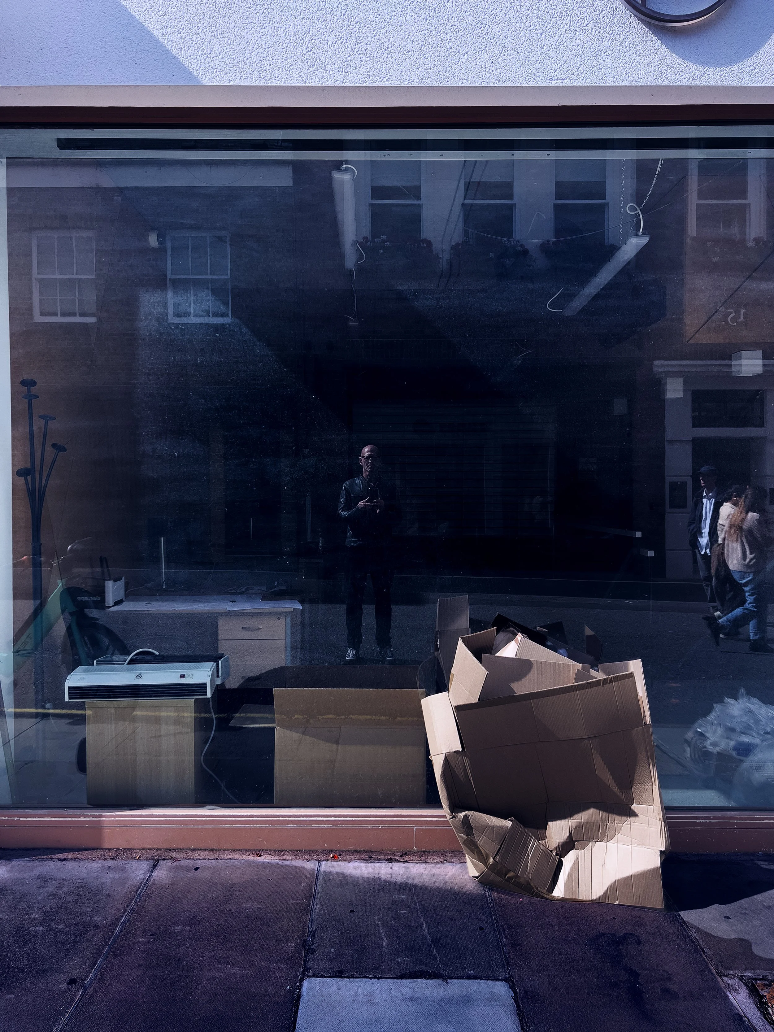 View through a large glass window showing reflections of buildings and people outside, a cardboard box on the ground outside, and an empty interior with a desk, computer, and furnishings inside.