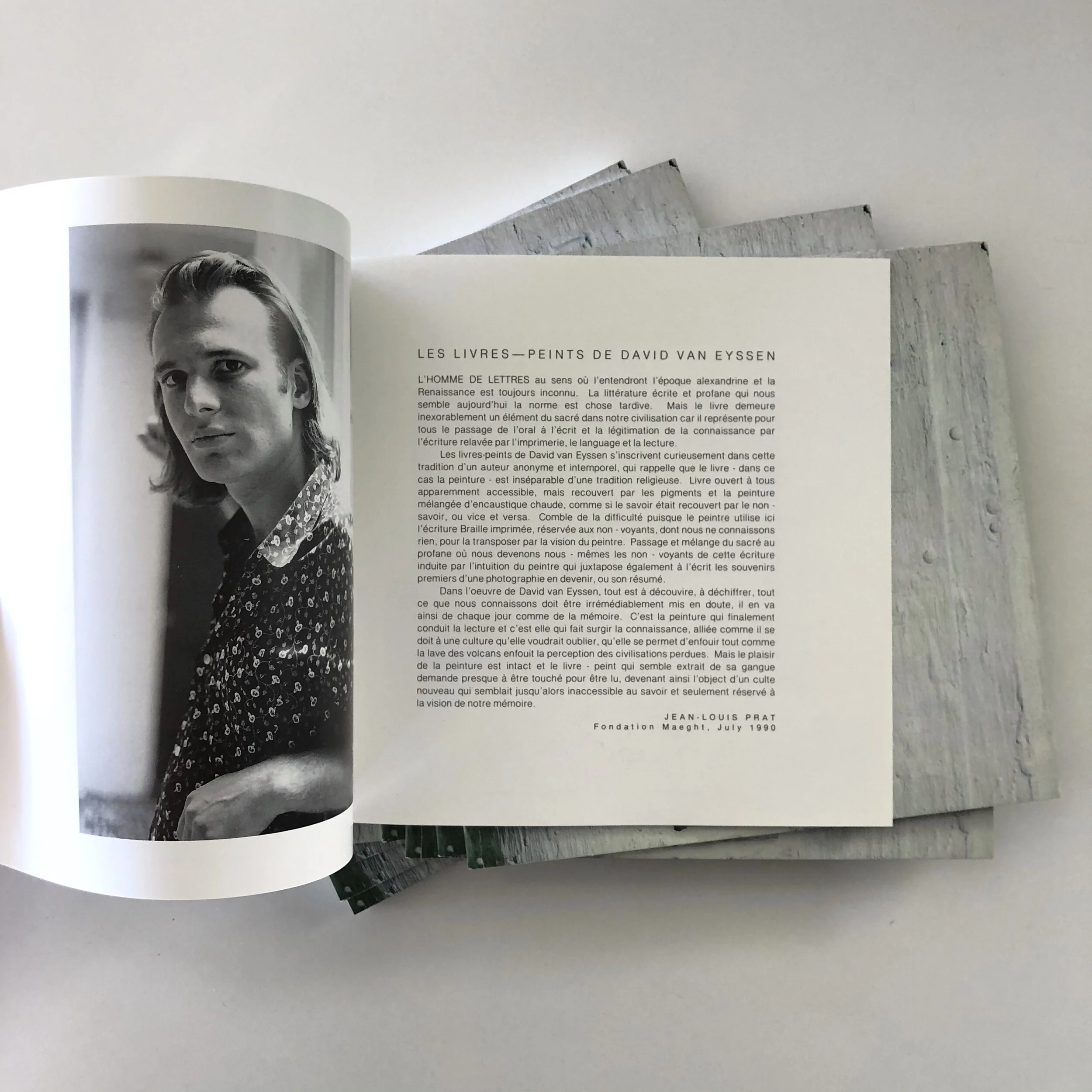 Open book showing a black and white photograph of a person and French text next to it. The page is decorated with a printed artwork overlap, with additional pages visible underneath.