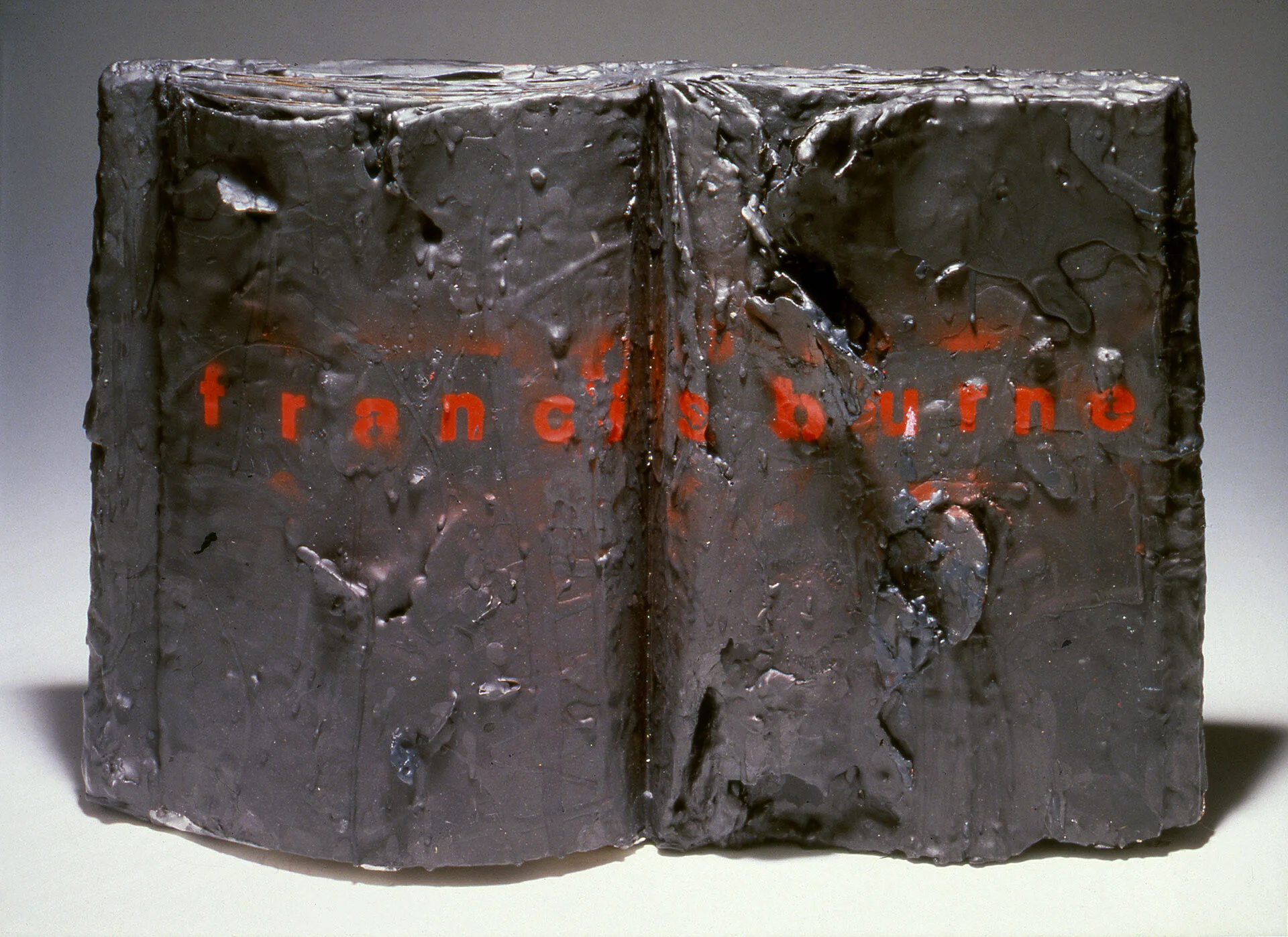 Artistic sculpture resembling a closed book with an abstract, textured surface and red text partially visible.