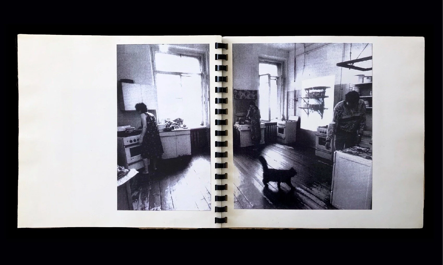 Black and white photograph of a kitchen. On the left page, a person in a dress is standing by a stove. On the right page, a person leans forward in another part of the kitchen, with a cat walking in the foreground. The images are printed in a book wi