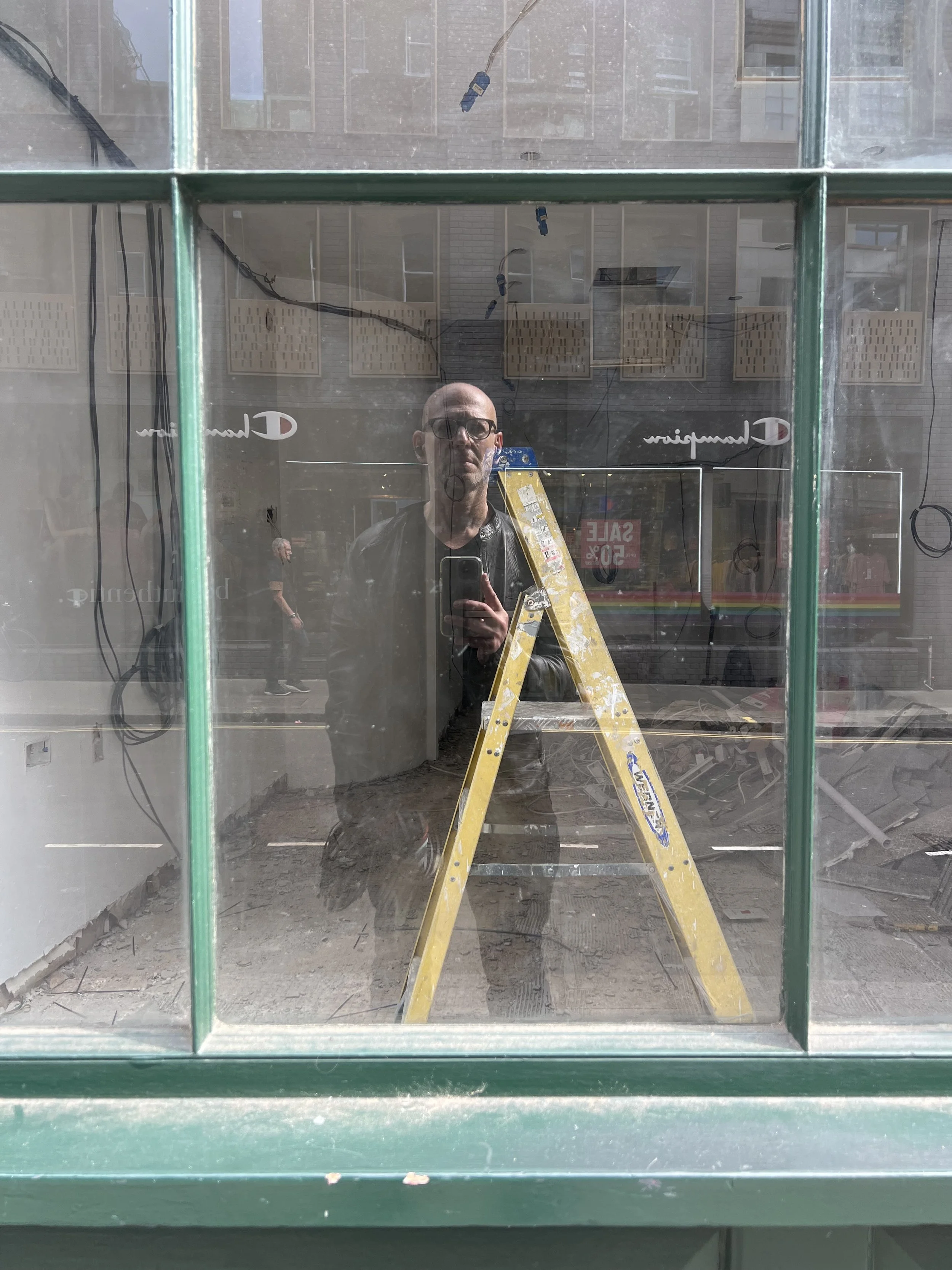 Reflection of a person taking a photo in a glass window, with an indoor scene showing a yellow ladder and construction debris.