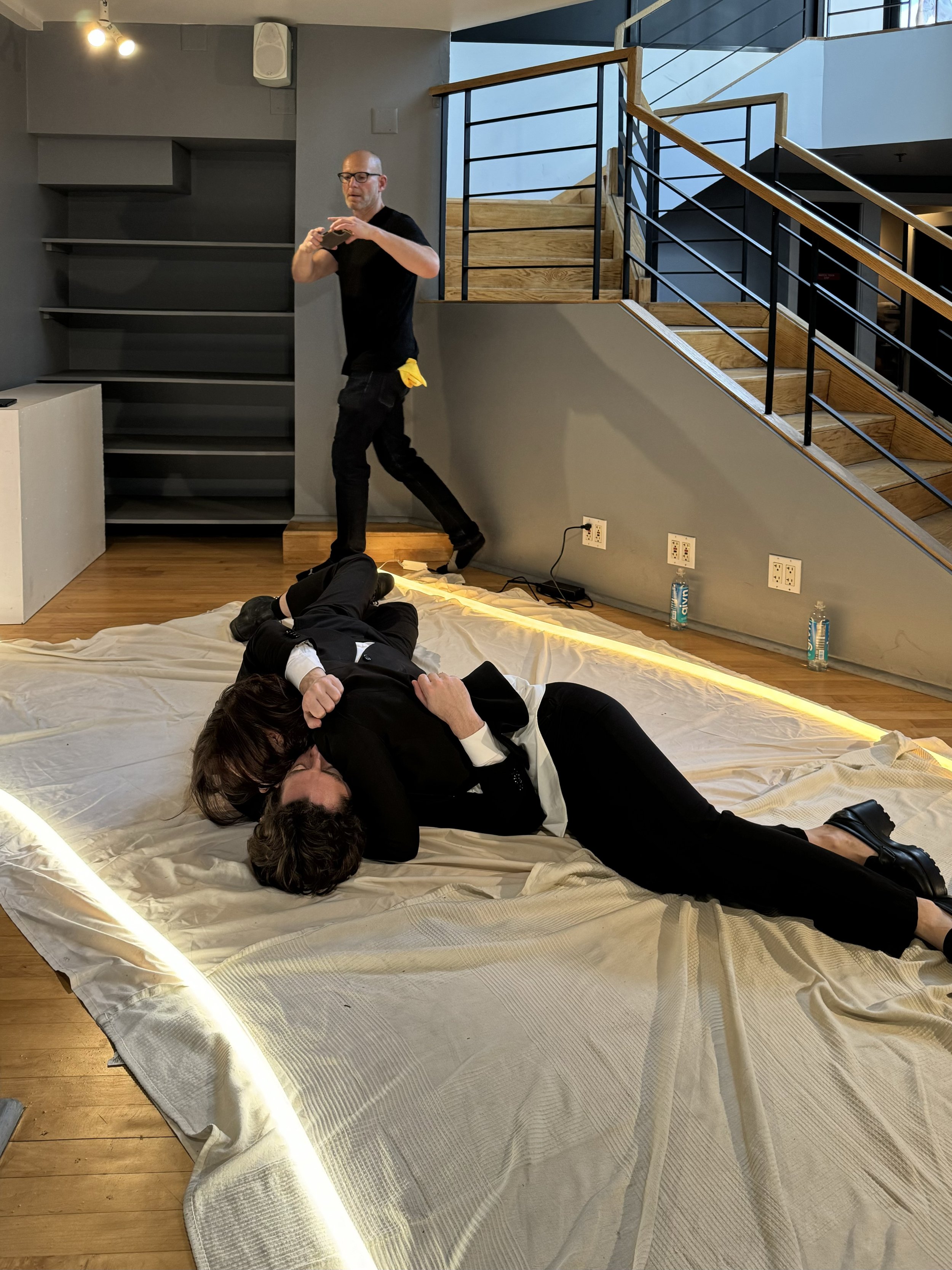 Two people lying on a beige blanket on the floor, embracing, with a man in the background gesturing near a staircase and shelves.