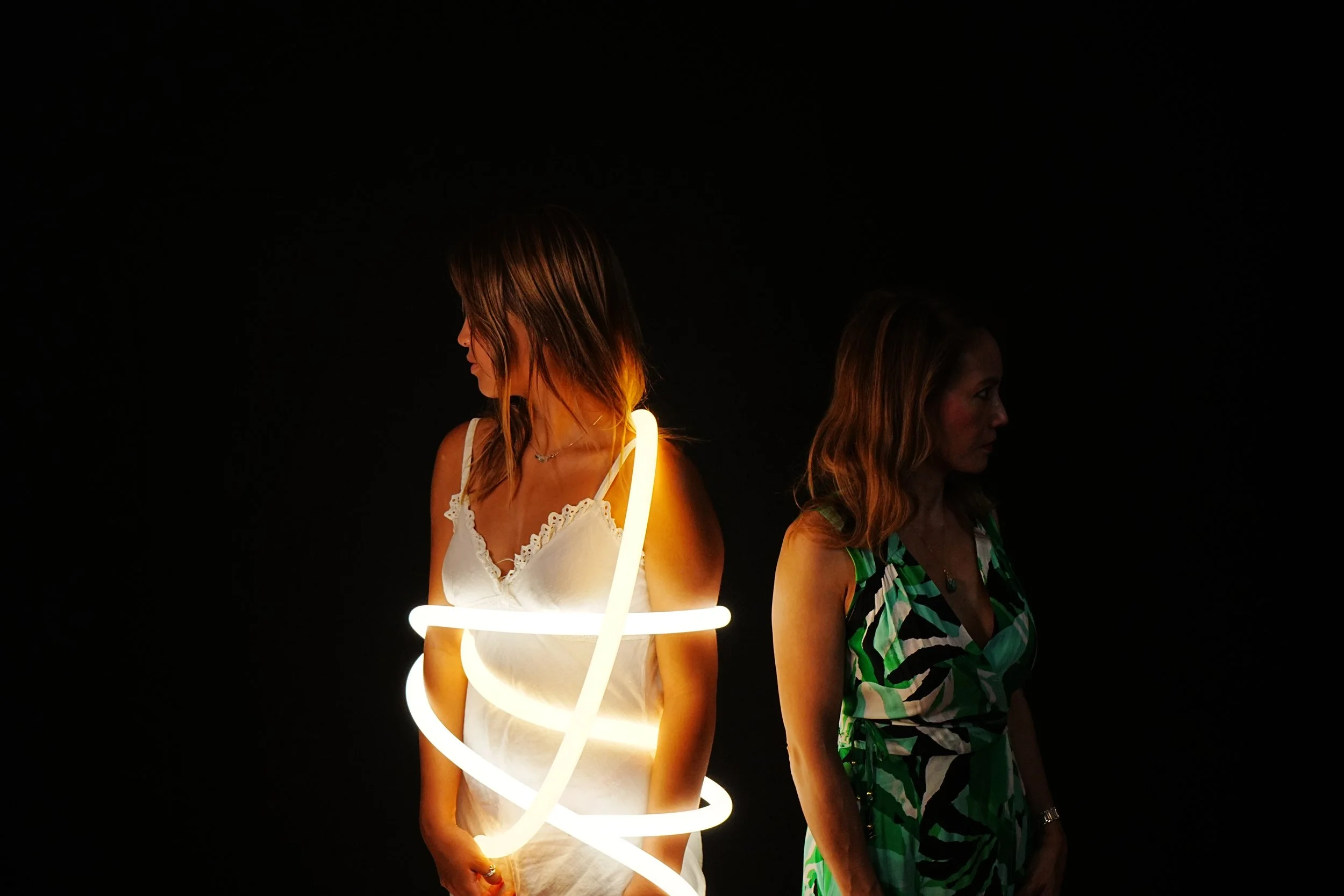 Two women standing back-to-back, one wrapped in glowing light strip, dark background.