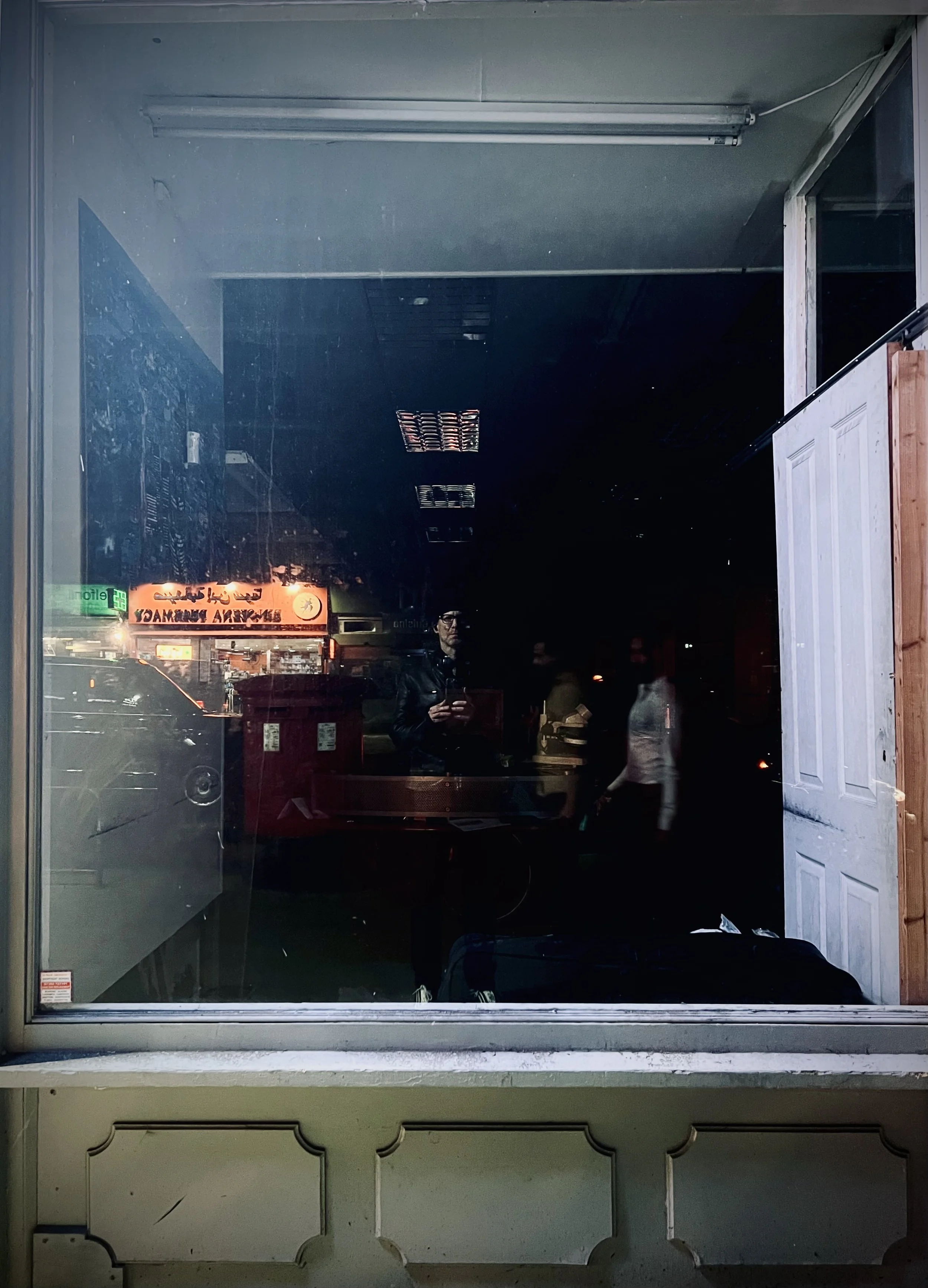 Reflection of a person in a glass window at night, with a lit sign in the background and a partially open door on the right.