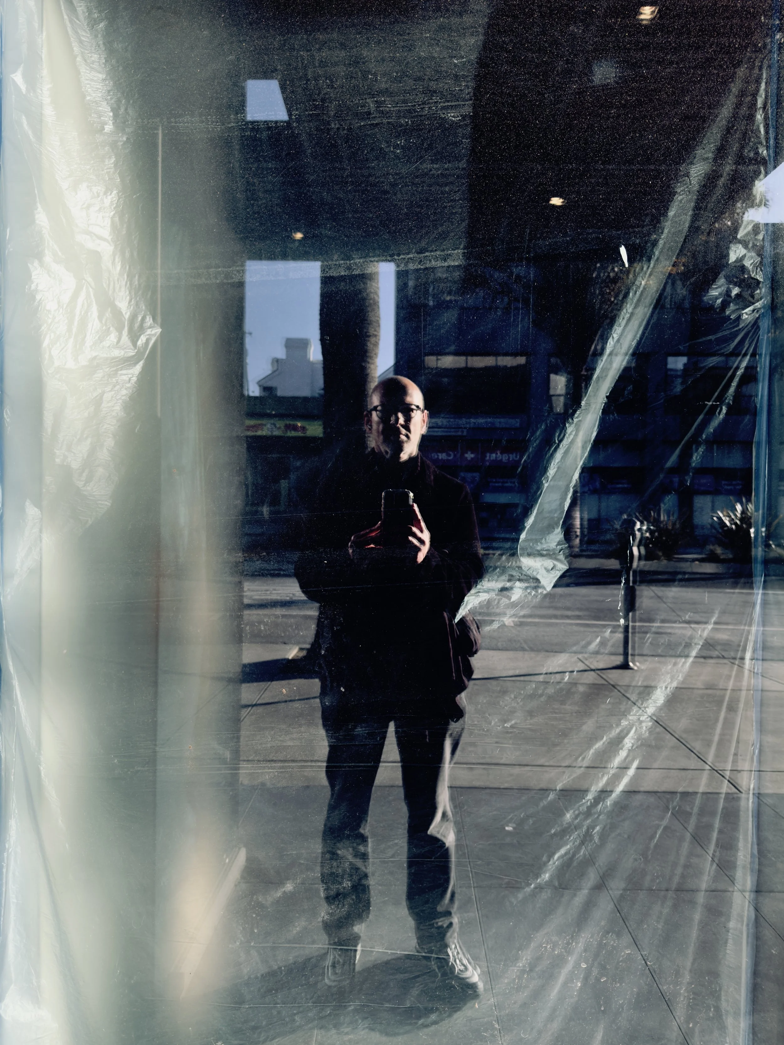 A person taking a selfie with a smartphone in front of a reflective surface, likely a large window or mirror, with urban buildings and street visible outside.