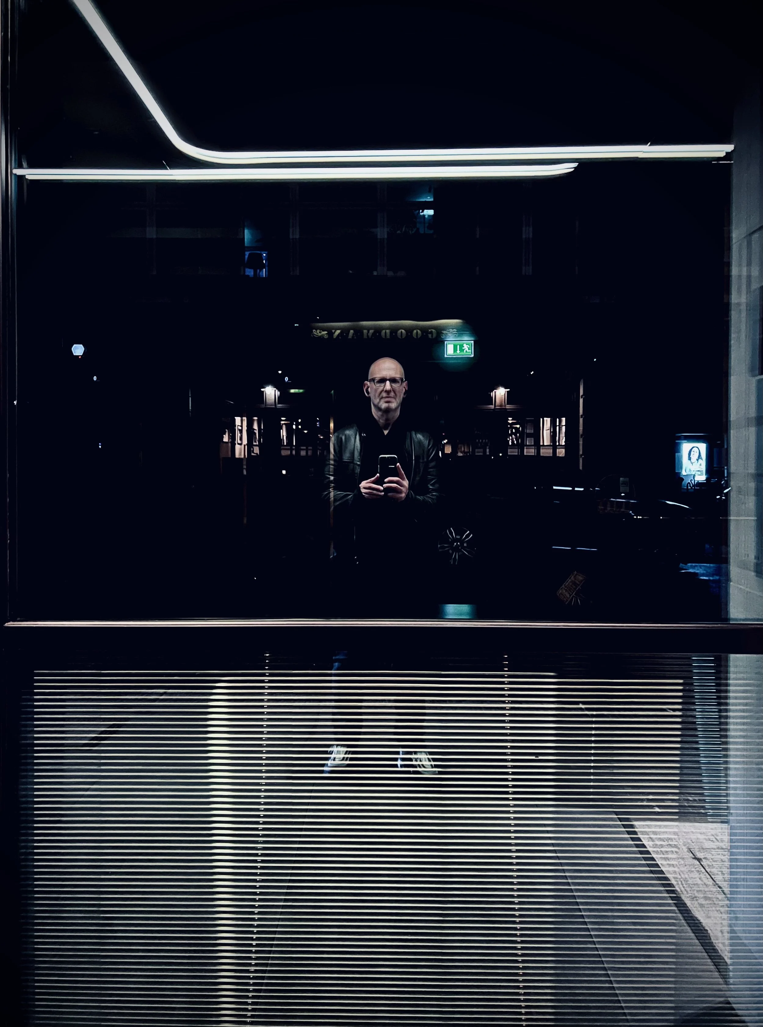 Reflection of a man taking a selfie in a dark glass window at night, with illuminated signs and street scene in the background.