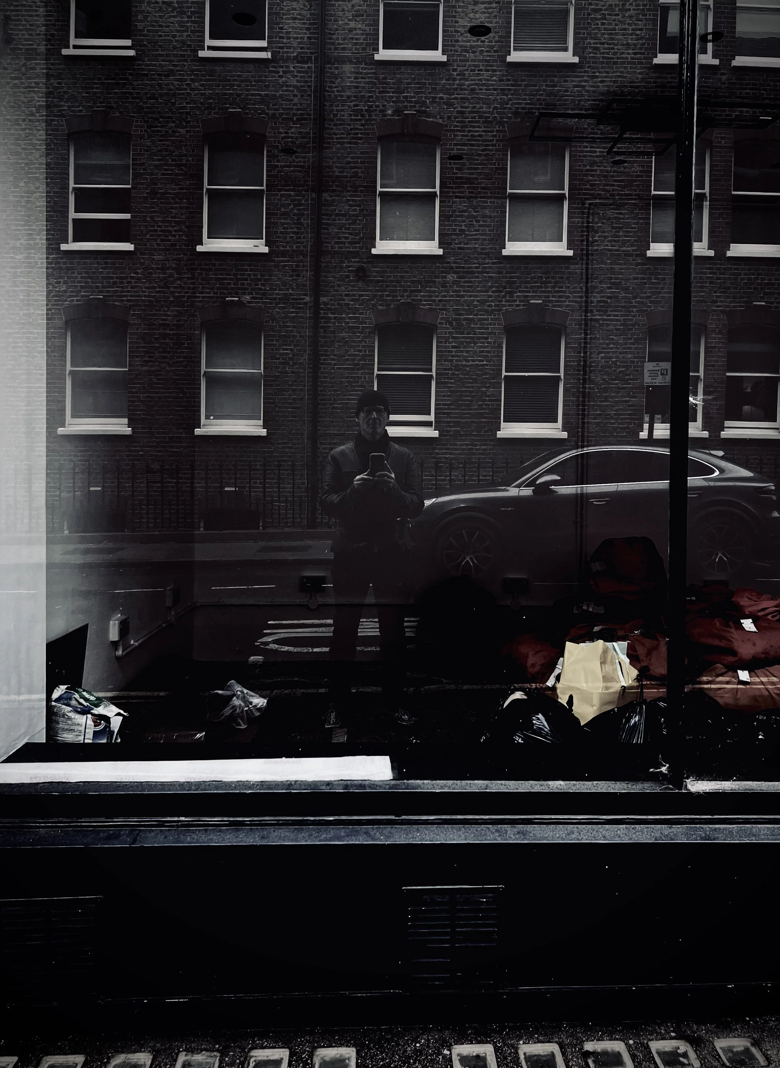 Reflection of a person taking a photo with a smartphone in a glass window. The background shows a brick building and parked car on a street.