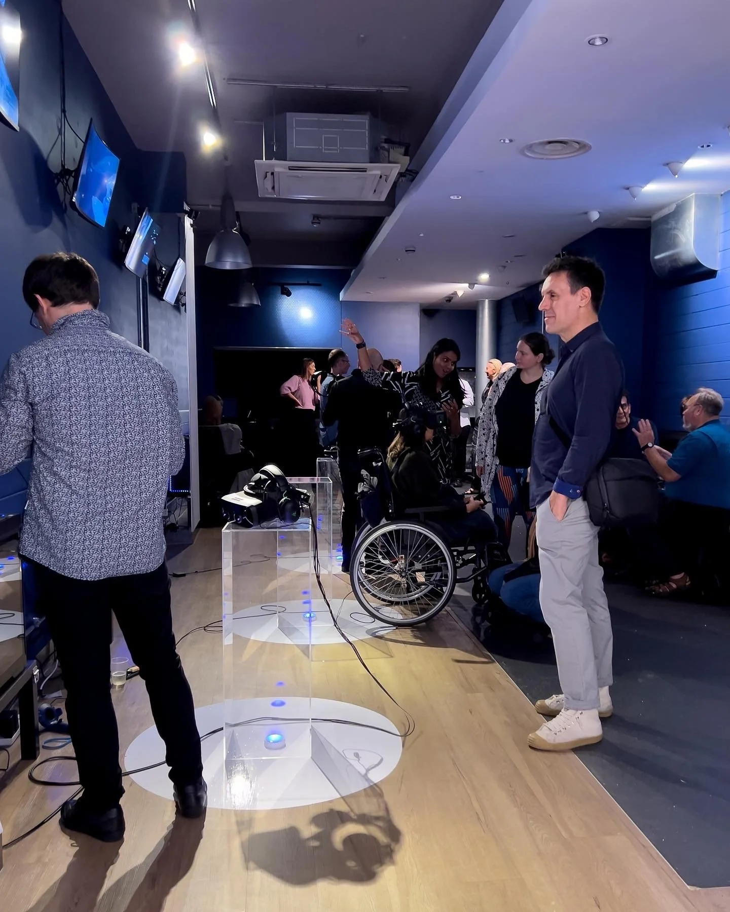 People gathered in a modern room with blue walls, some interacting and others observing. A person in a wheelchair is present, and virtual reality headsets are visible on a transparent stand. The atmosphere is social and semi-casual.
