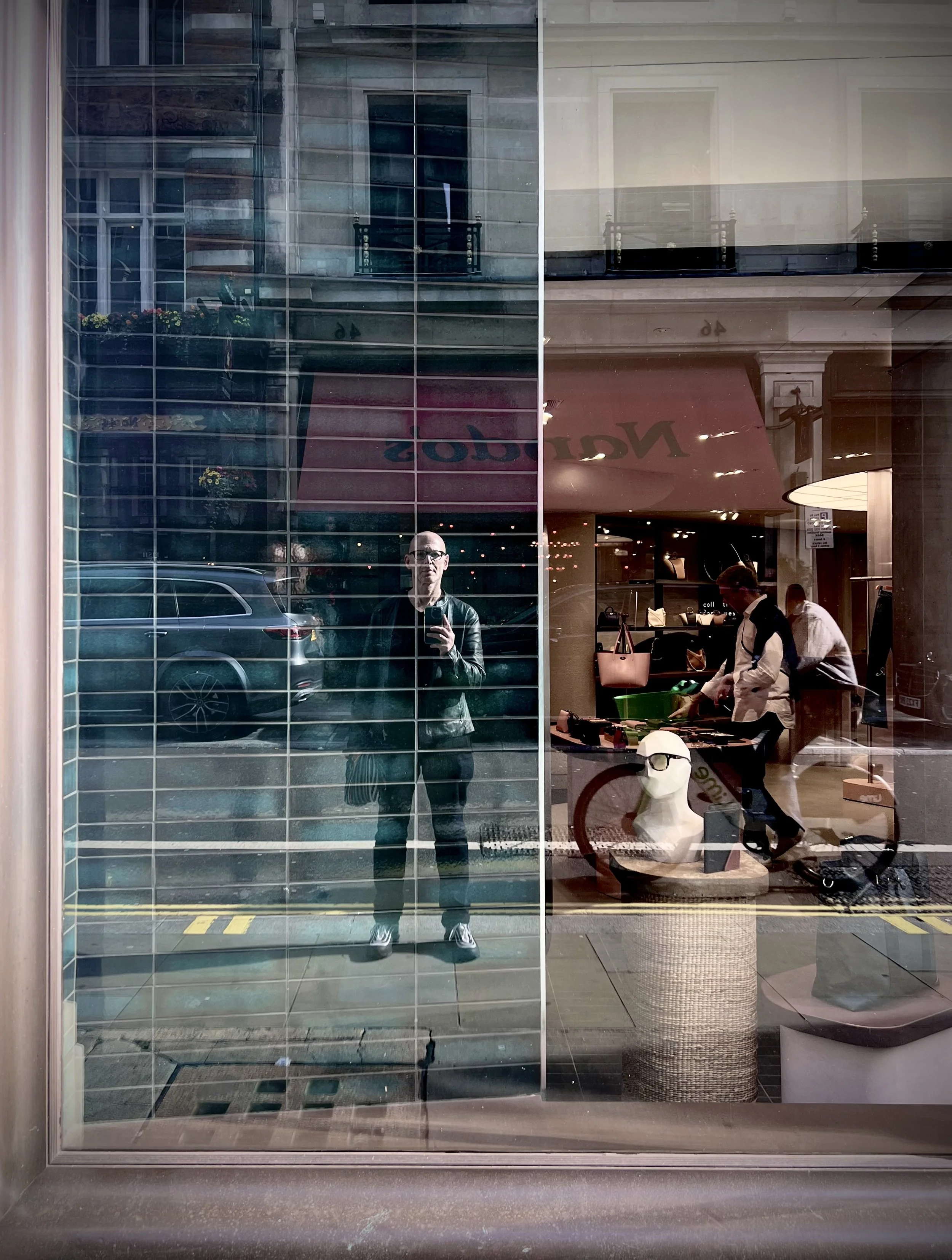 Reflections in a shop window showing a man taking a photo, a person with a shopping cart, mannequins with sunglasses, and buildings across the street.
