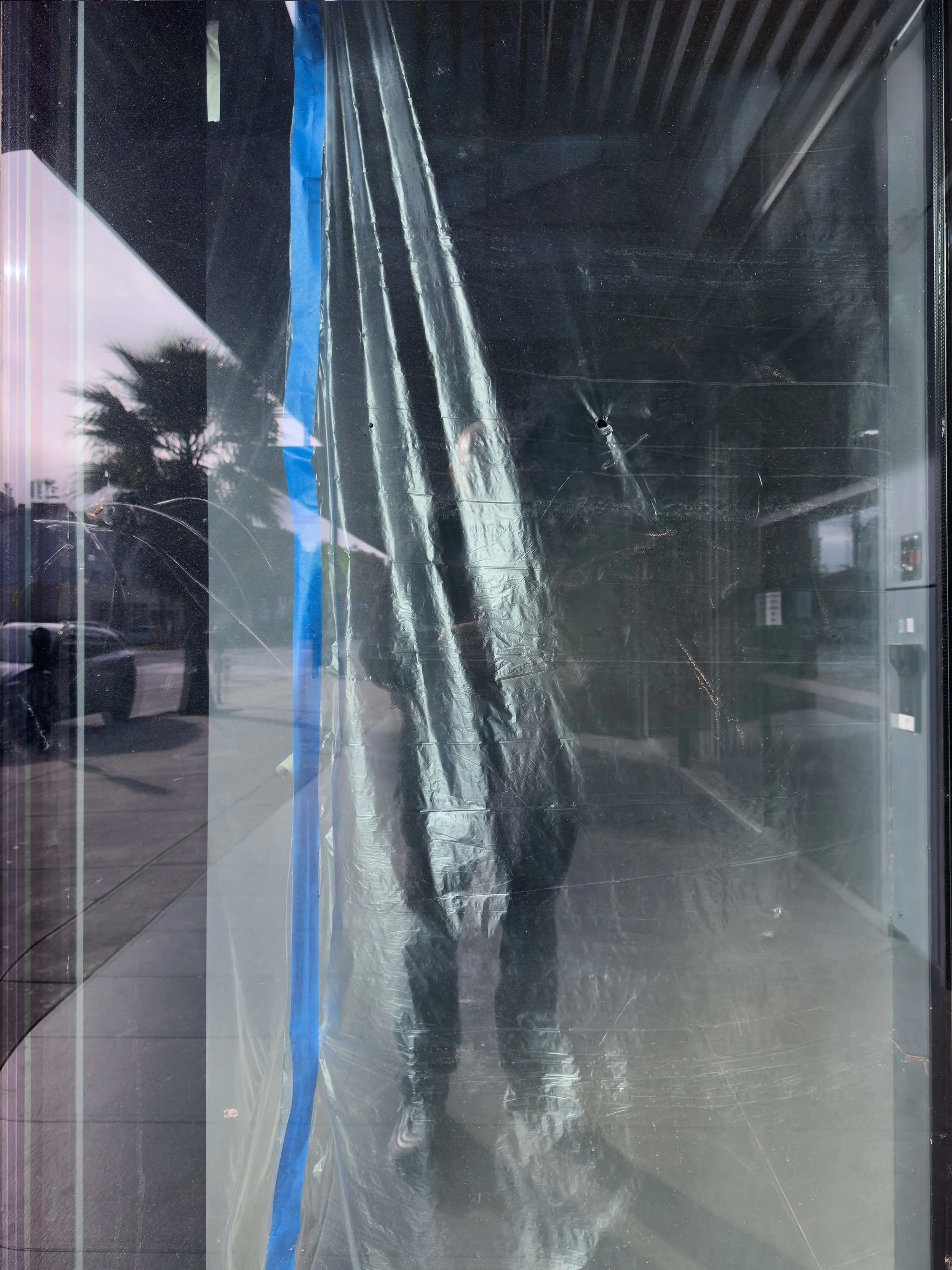 Photo of a person taking a picture of their reflection in a window that has protective plastic sheet and tape on it, with a palm tree and street scene reflected in the glass.