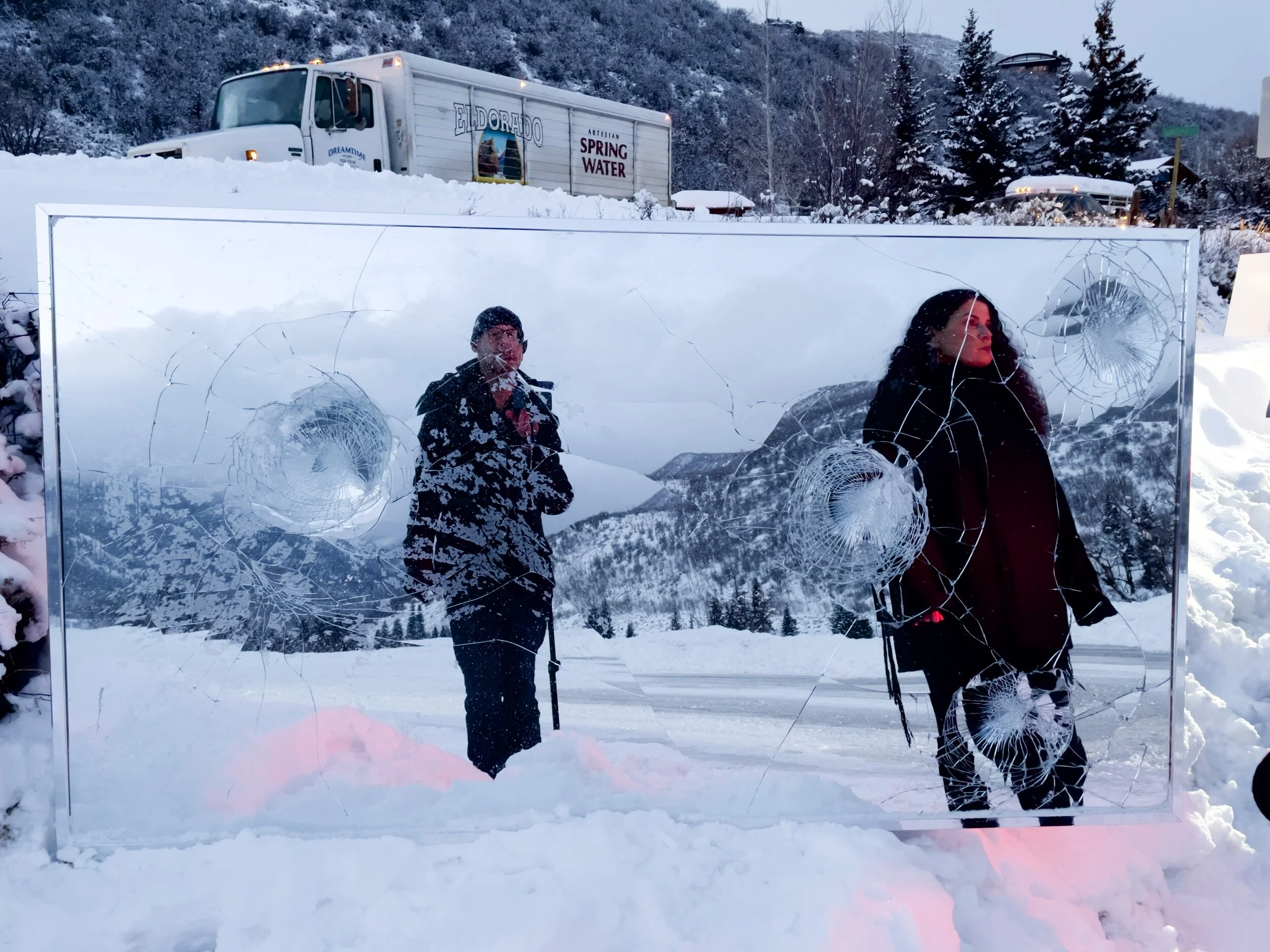 Two people reflected in a broken mirror amidst snow, with a snow-covered landscape, trees, and a truck labeled "Eldorado Spring Water" in the background.