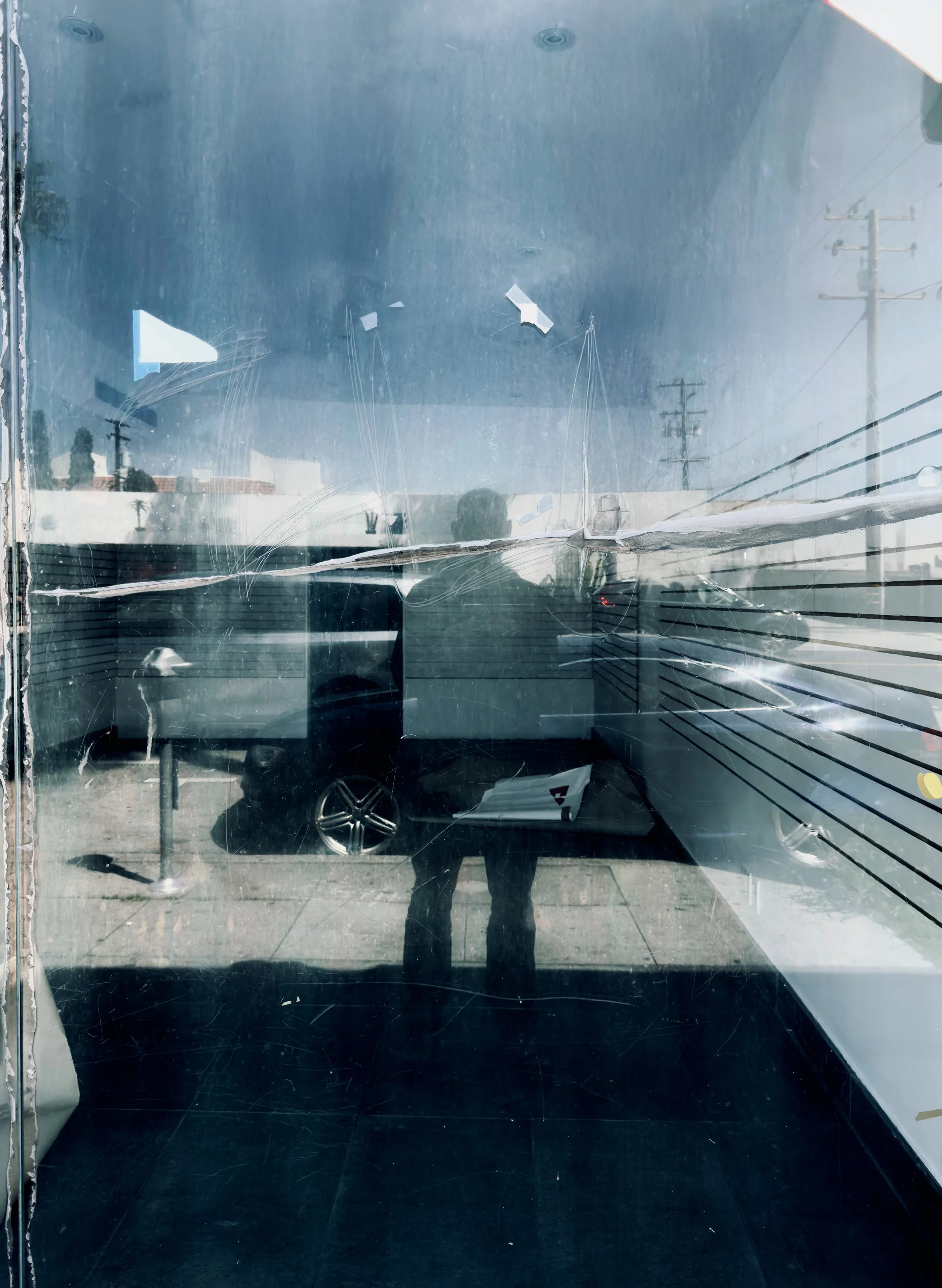 The image shows a front window that is scratched and has small torn pieces of paper or stickers on it. The reflection shows a parked SUV, a person taking the photo, and utility poles against a blue sky with some clouds.