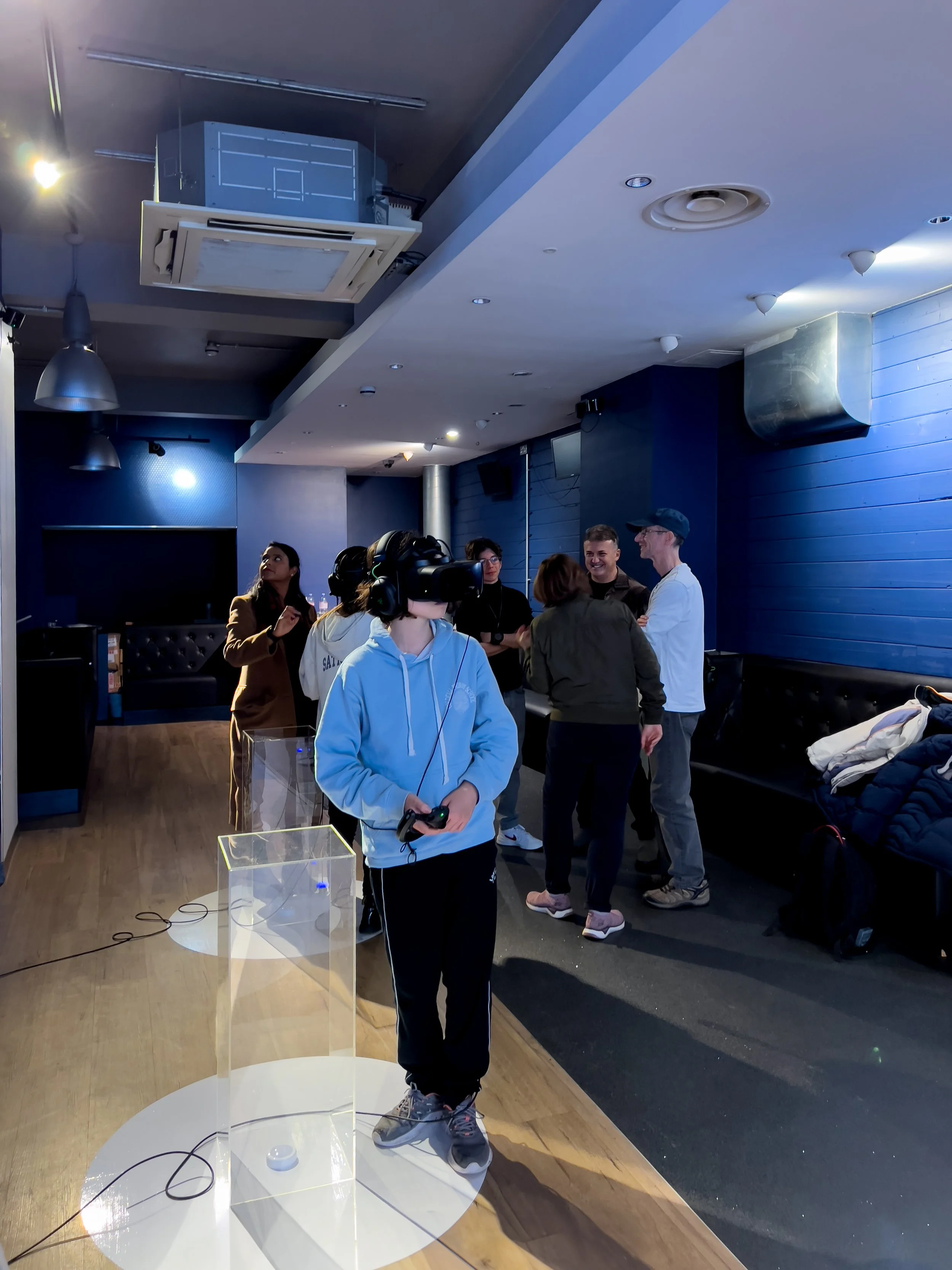 A person wearing a blue hoodie and VR headset stands in a modern room with wooden flooring and blue walls. They hold a controller. Several people in the background are conversing. The room has ceiling lights and metal ductwork, and coats are piled on