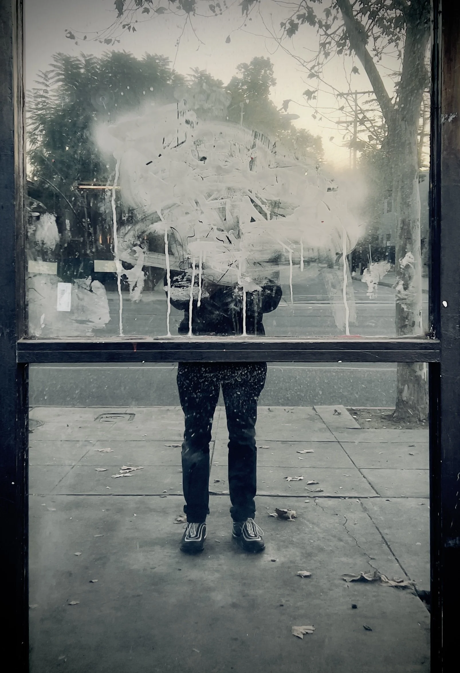 Reflection of a person taking a photo in a dirty window with smudges and streaks, outside street scene with trees and pedestrians.