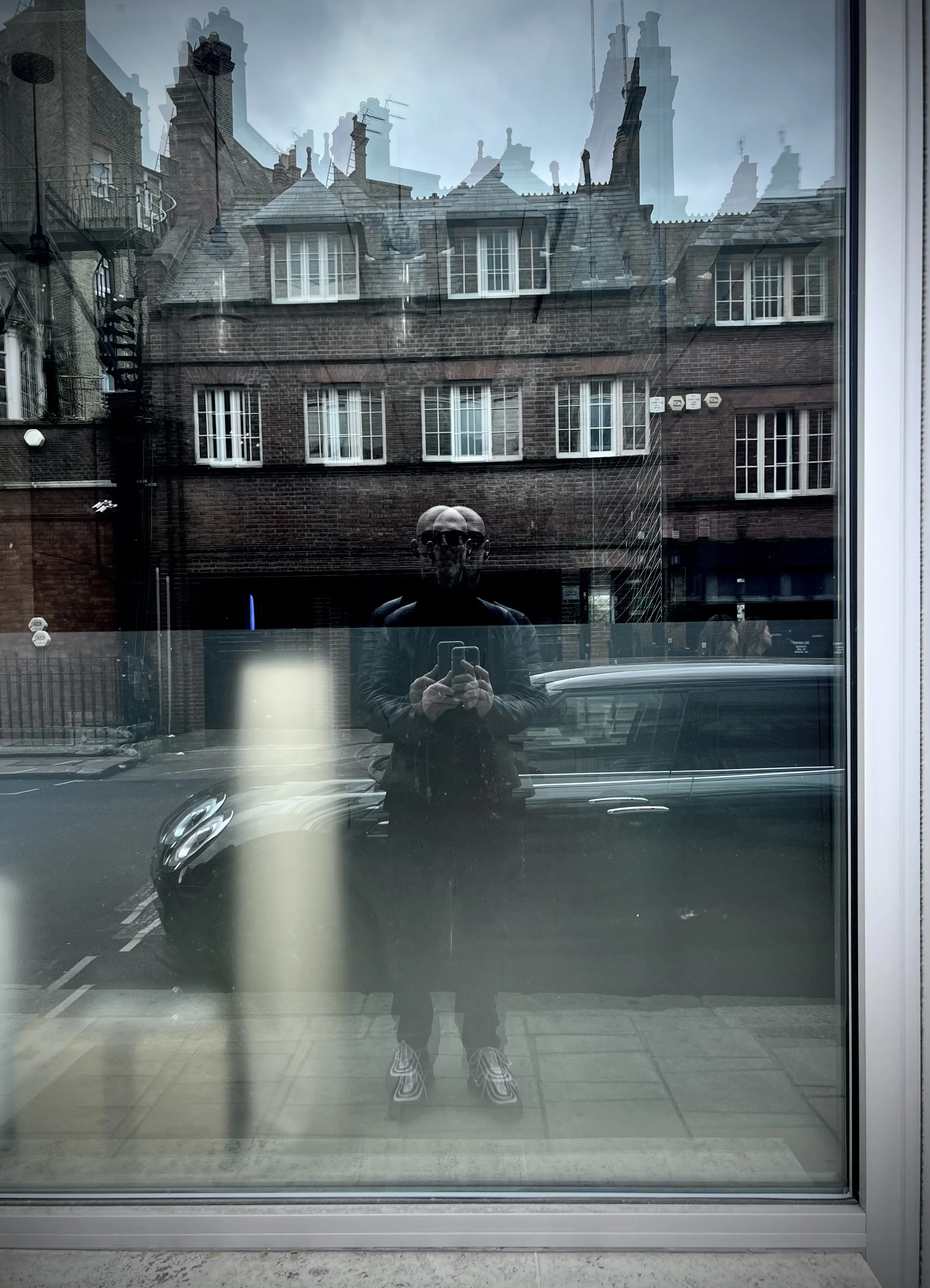 Reflection of a person taking a photo in a glass window, with old brick buildings and cars visible through the glass.