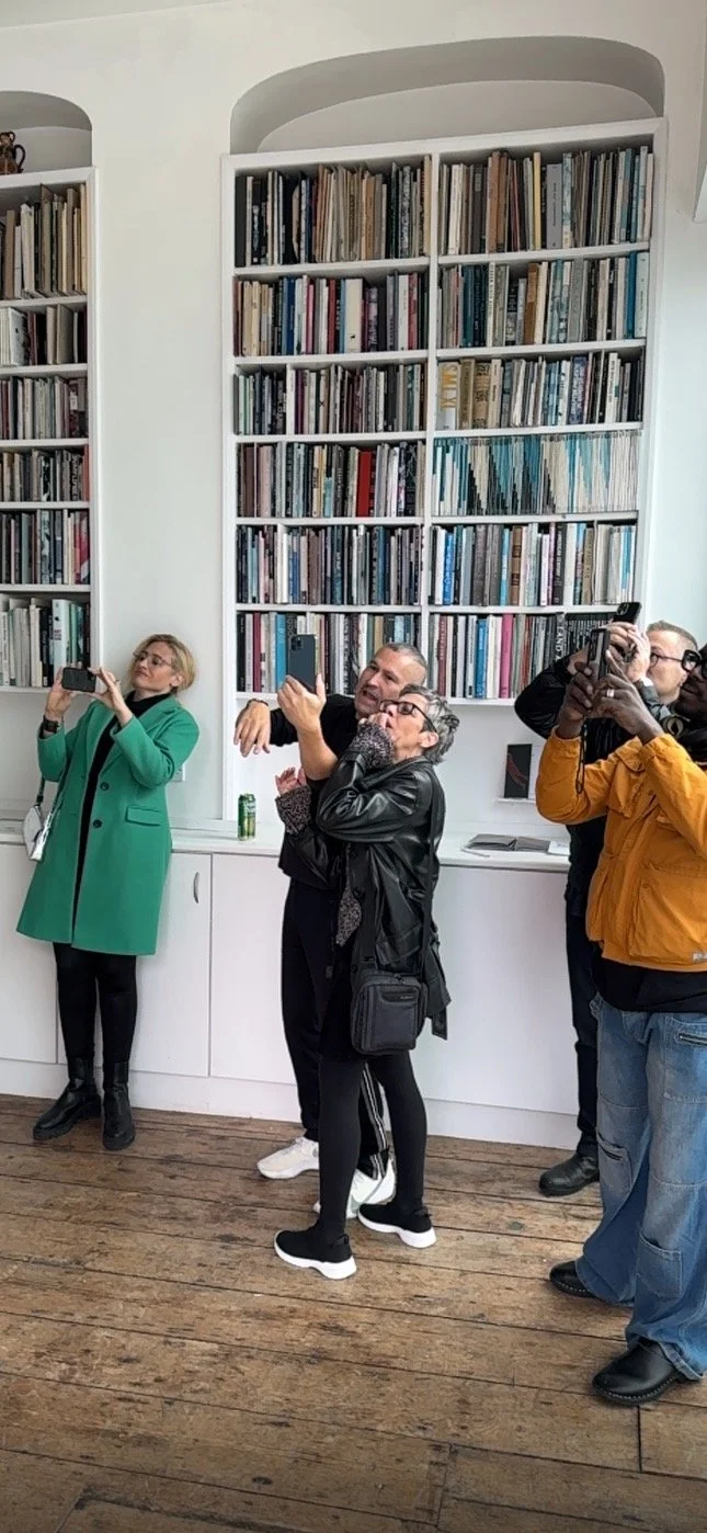 A group of people taking photos with their smartphones in front of a wall of bookshelves filled with books. They are standing on a wooden floor in a modern, bright room.