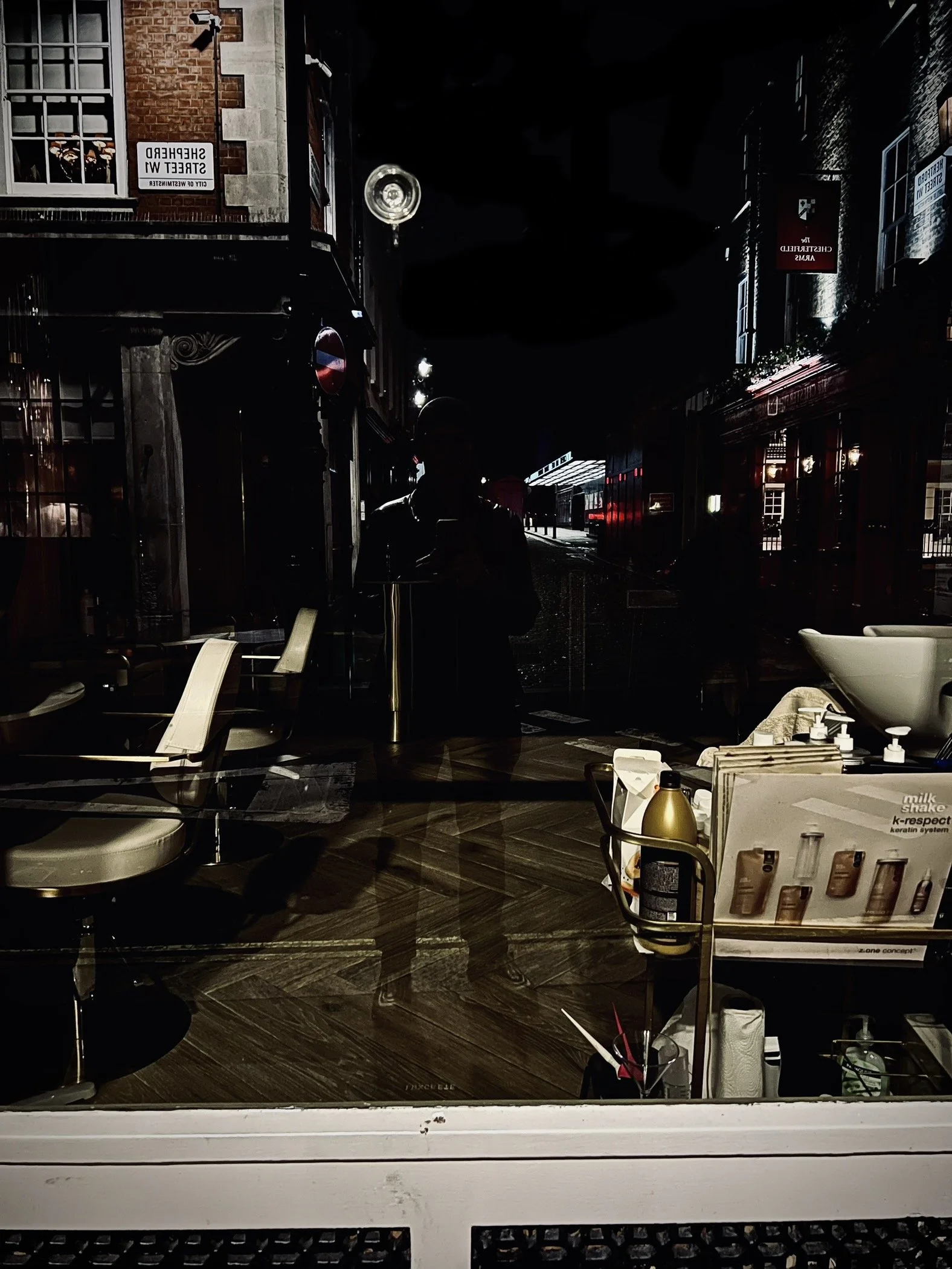 Nighttime view through a restaurant window showing chairs and tables indoors, a person taking a photo, and reflections of street signs and buildings outside.