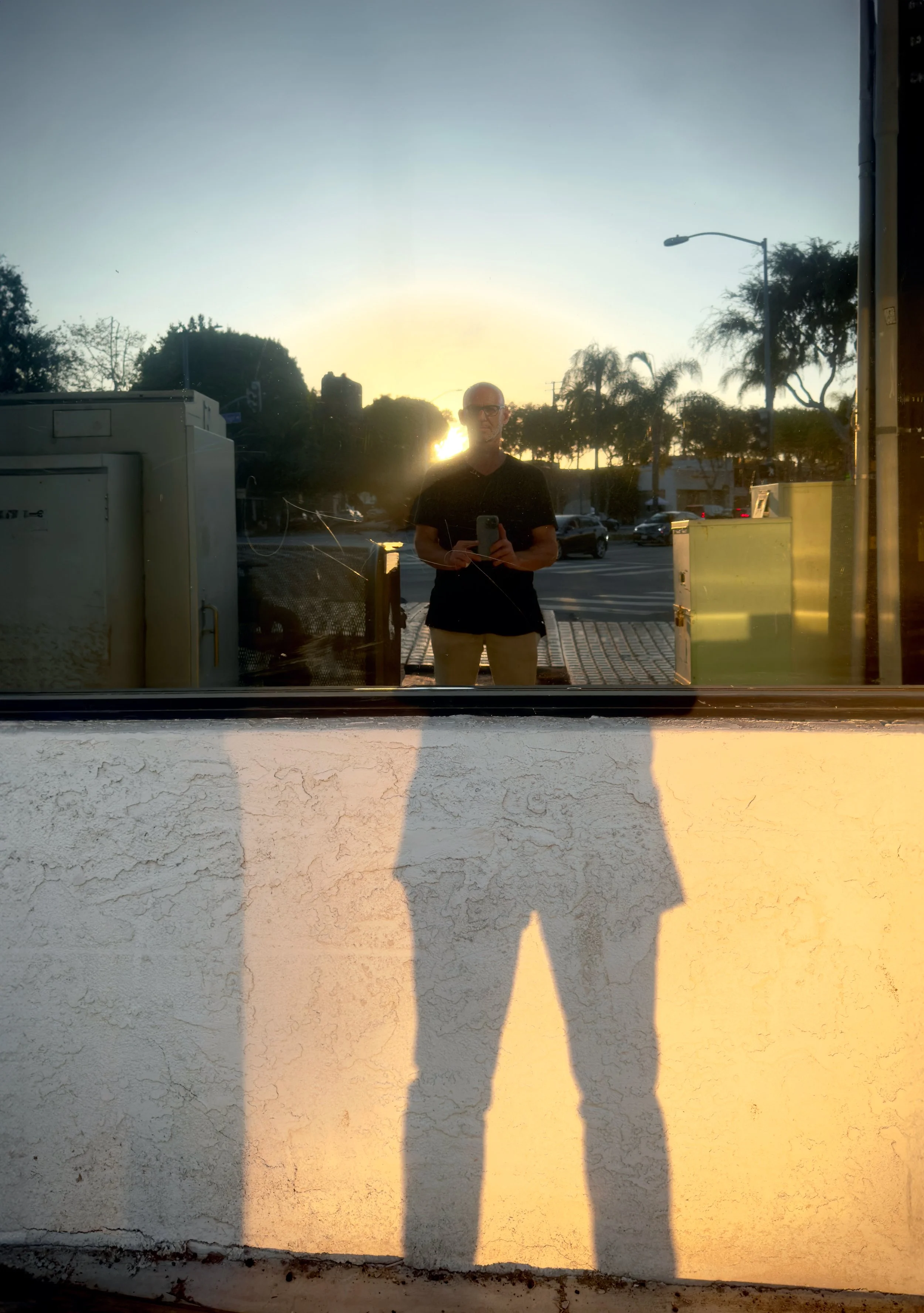 Reflection of a man holding a phone in front of a window, with the sunset behind him, casting a shadow on the wall below.