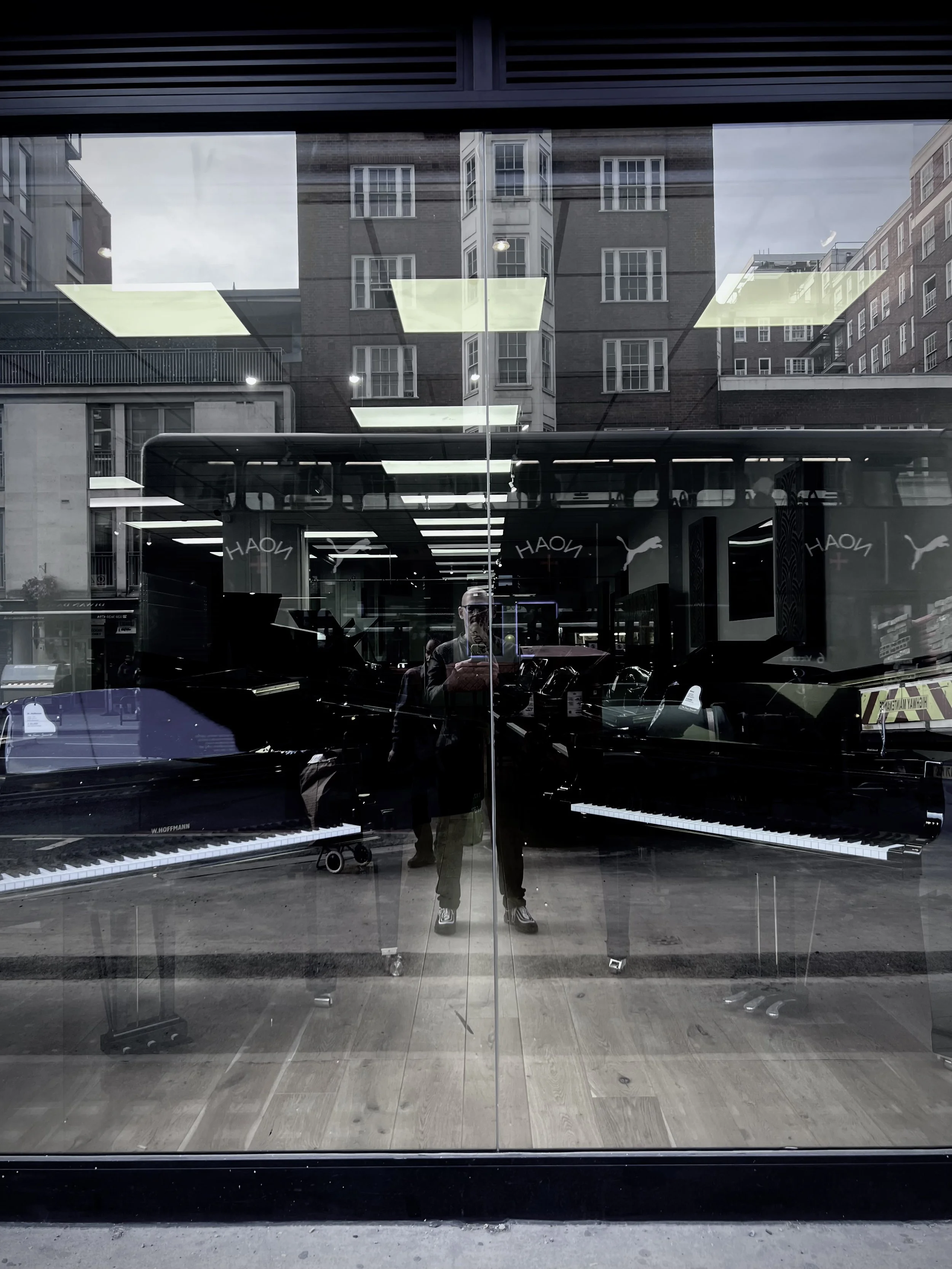 Person photographing through glass window of music store with reflections of buildings, displaying pianos inside.